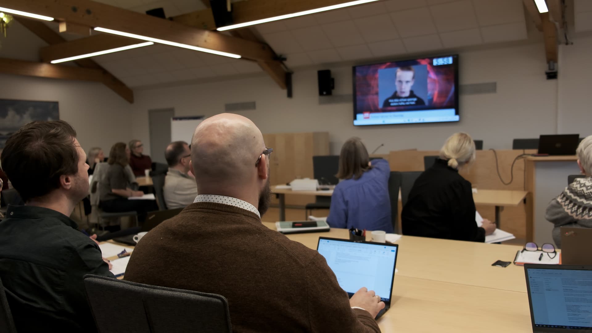 I et konferanserom sitter det folk og følger med på en presentasjon på en storskjerm som viser en mann som taler. Noen av deltakerne har bærbare datamaskiner åpne og tar notater. Rommet har trebjelker og moderne belysning.