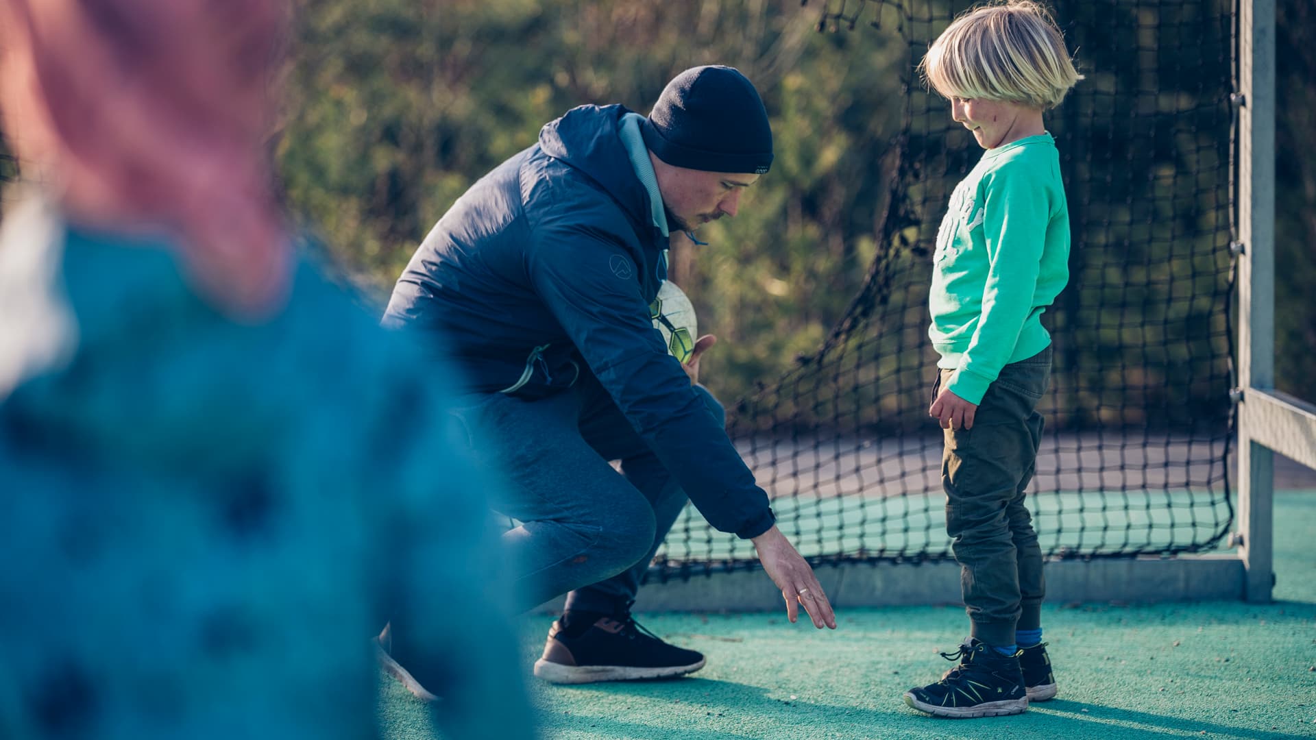 En mann står på kne på en idrettsbane og gestikulerer mot bakken mens han snakker med et lite barn som står foran ham. En annen uskarp skikkelse er synlig i forgrunnen. Et nett er i bakgrunnen.