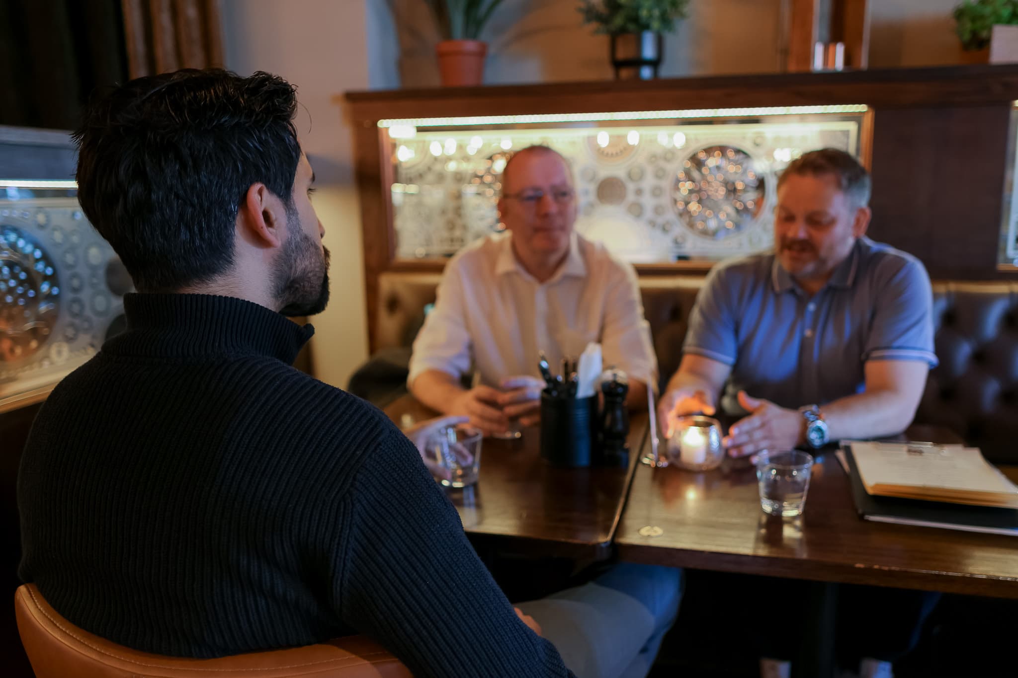 Tre menn sitter ved et restaurantbord og snakker sammen. To av mennene er vendt mot kameraet, mens den ene sitter med ryggen til. På bordet står det drinker, et stearinlys og bestikk. Settingen virker uformell og avslappet.
