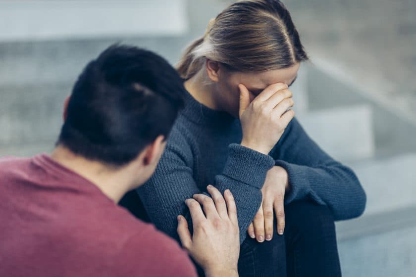 En person sitter på en trapp, dekker ansiktet med den ene hånden og ser opprørt ut, mens en annen person forsiktig berører armen deres i en trøstende gest.