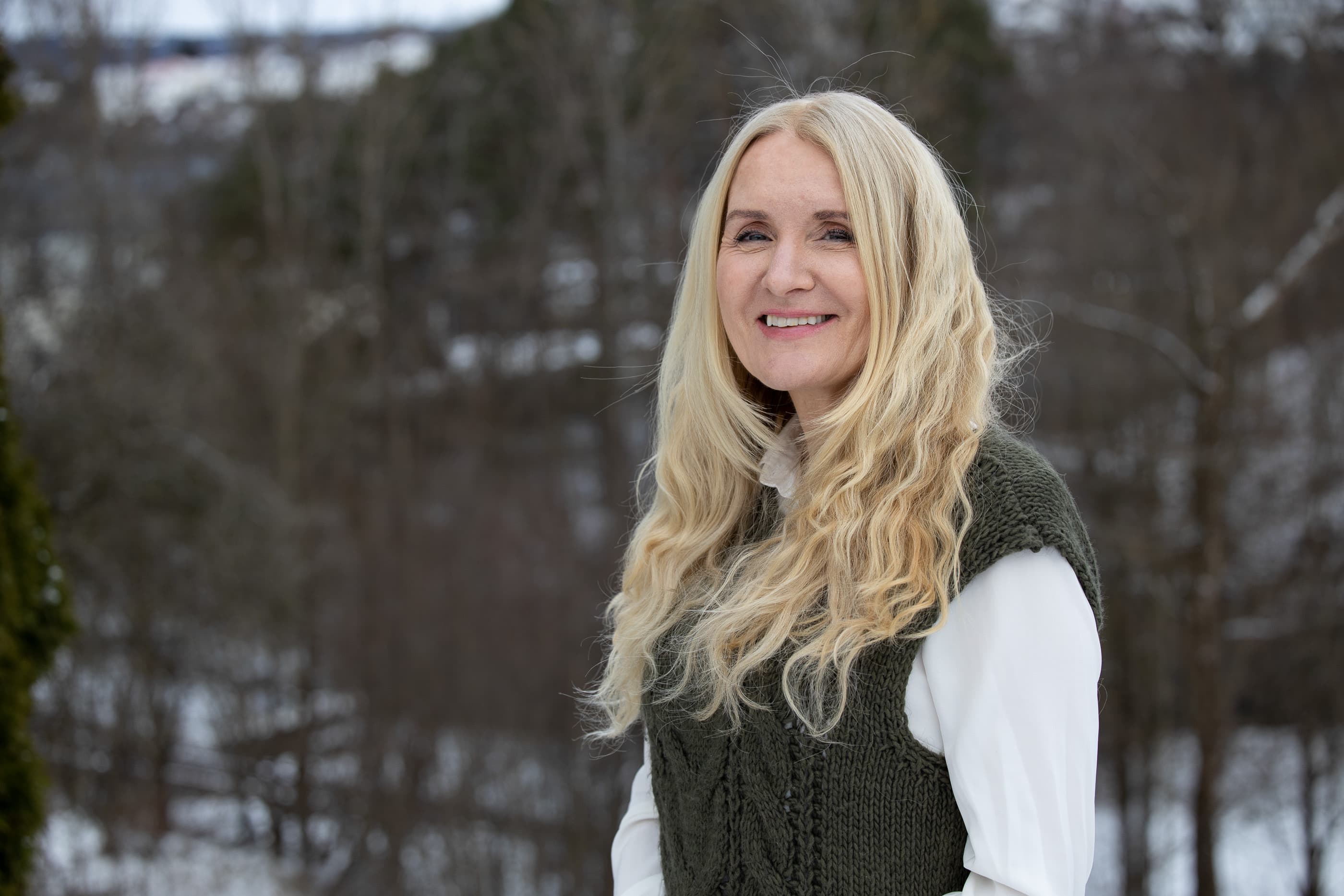 En smilende kvinne med langt, blondt hår, hvit skjorte og grønn strikkevest står utendørs i et snødekt skogsområde.