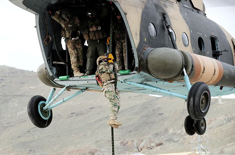 En soldat i kamuflasjetrekk rappellerer fra et militærhelikopter mens andre venter inne i helikopteret. Helikopteret svever over et steinete terreng med fjell i bakgrunnen.