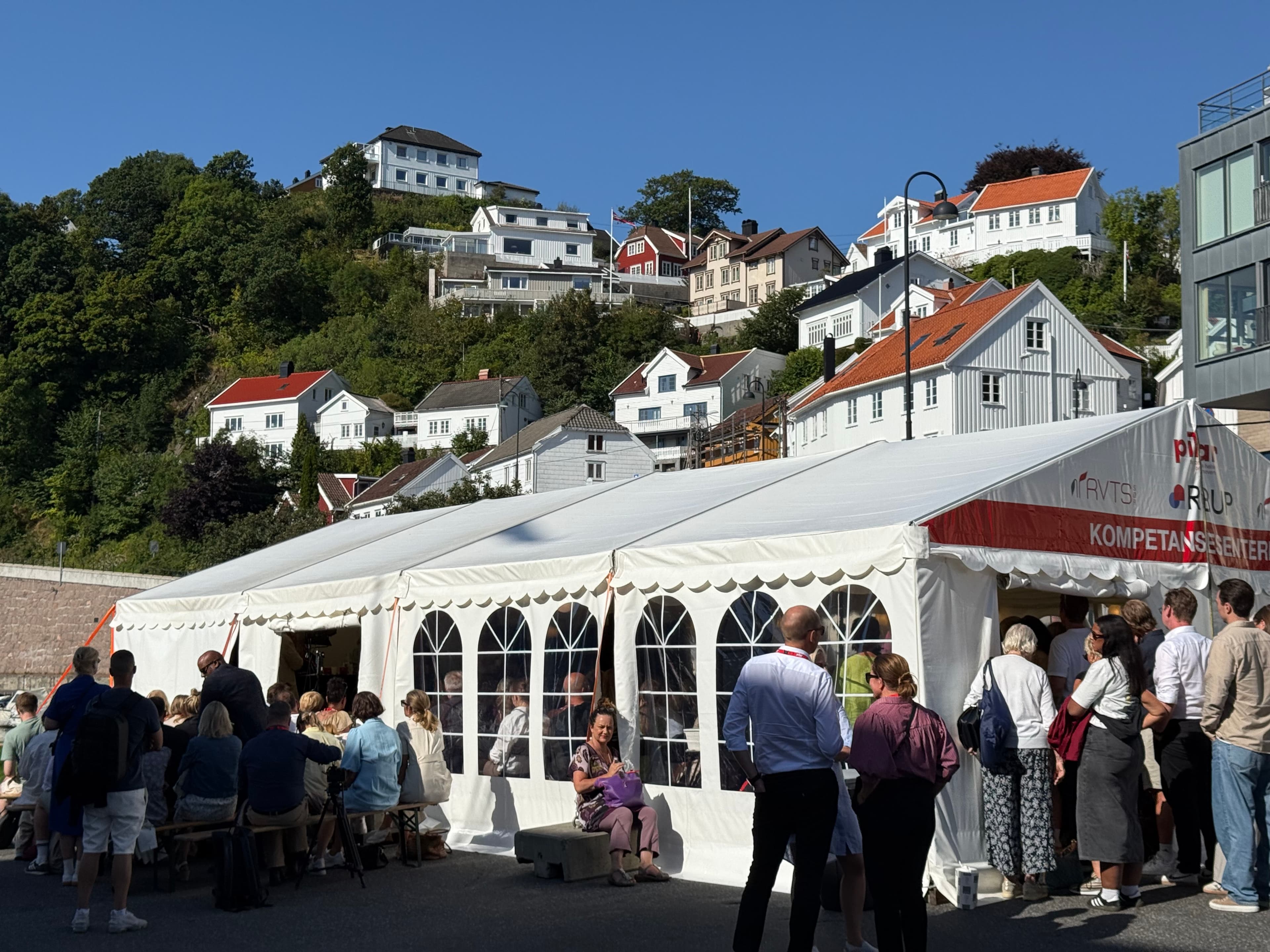 En folkemengde samler seg utenfor og inne i et stort hvitt festtelt på en solrik dag, med hvite trehus på en åsside og frodig grøntområder i bakgrunnen.