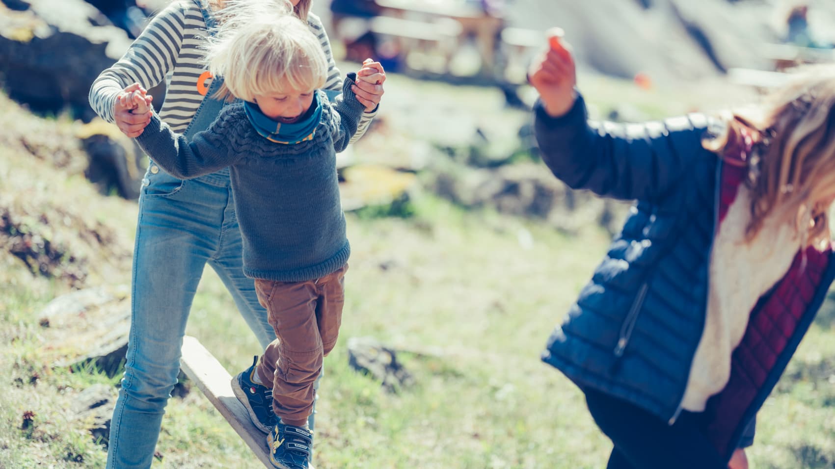 Et lite barn med blondt hår balanserer på en trebjelke utendørs og holder en voksen i hånden. I forgrunnen står en annen person i blå jakke. Omgivelsene er solfylte og gressbevokste.
