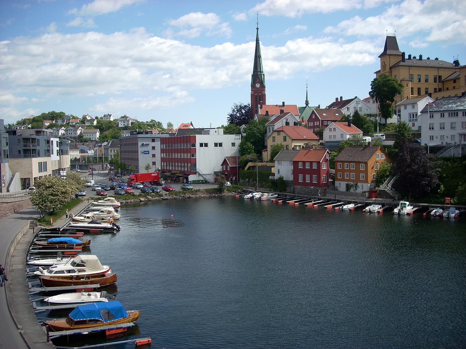 En liten havn med fargerike båter og hus langs vannet, med en kirke med et høyt kirketårn og andre bygninger i et kupert landskap under en delvis skyet himmel.