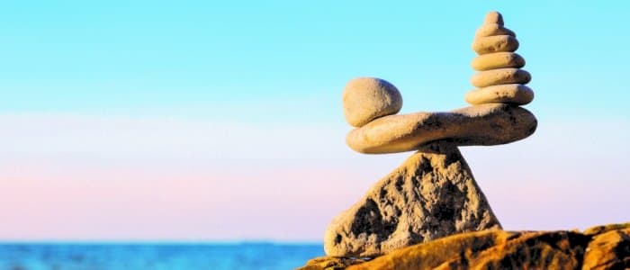 En stabel med glatte steiner balansert forsiktig på en stor stein ved sjøen, med en klar blå himmel og rolig vann i bakgrunnen.