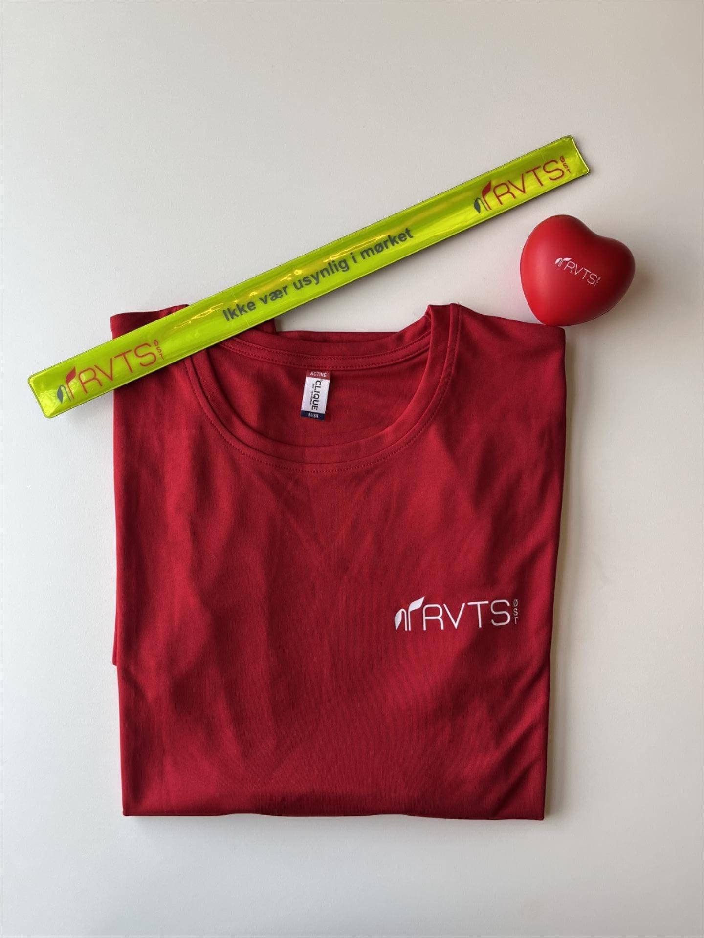 En rød brettet T-skjorte, et gult refleksarmbånd og en rød hjerteformet stressball, alle merket med RVTS-logoen, er plassert på en hvit flate.