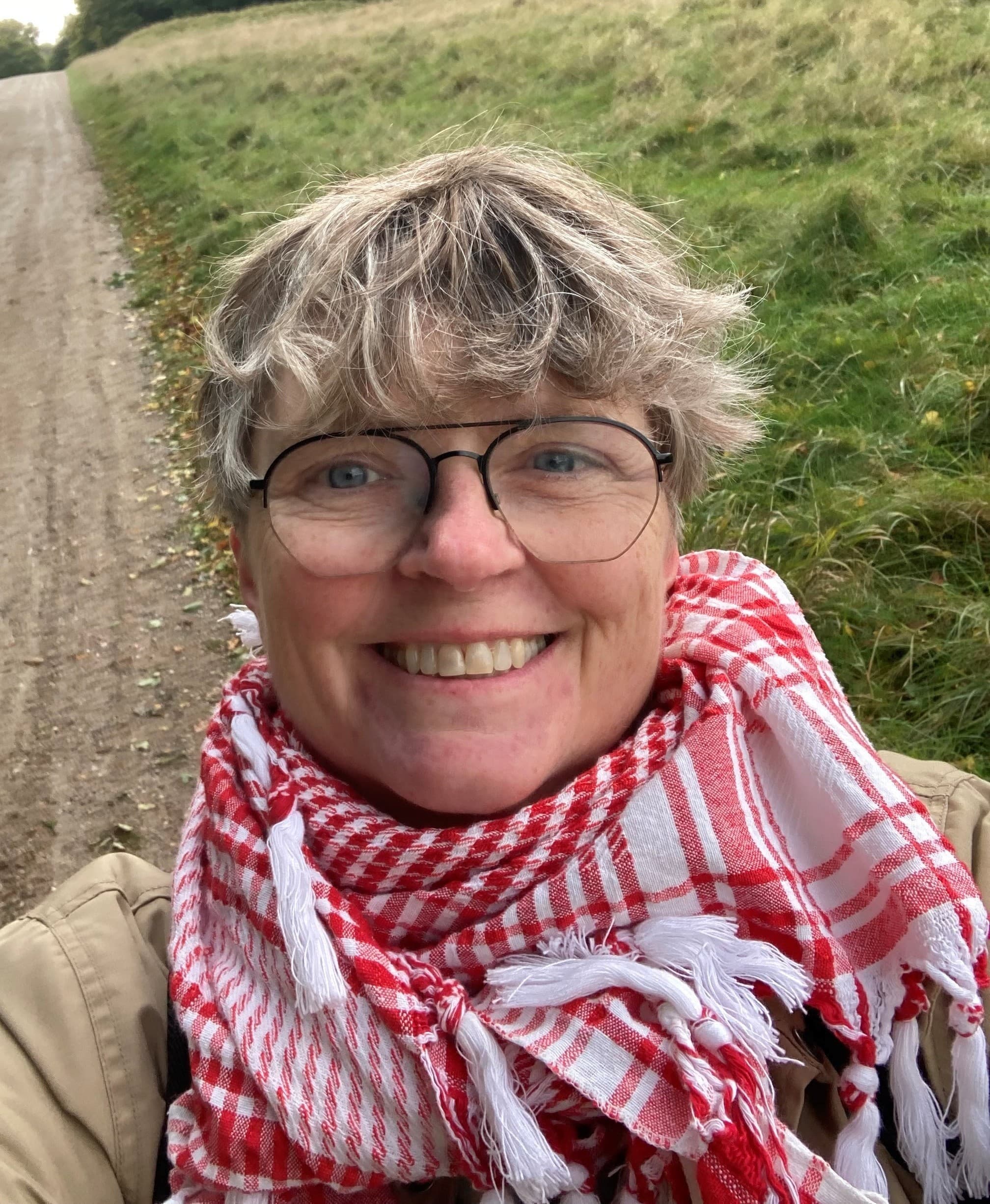 En person med kort, grått hår og briller smiler til kameraet mens han står ved siden av en gressbelagt sti, iført et rød- og hvitrutete skjerf og en brunbrun jakke.
