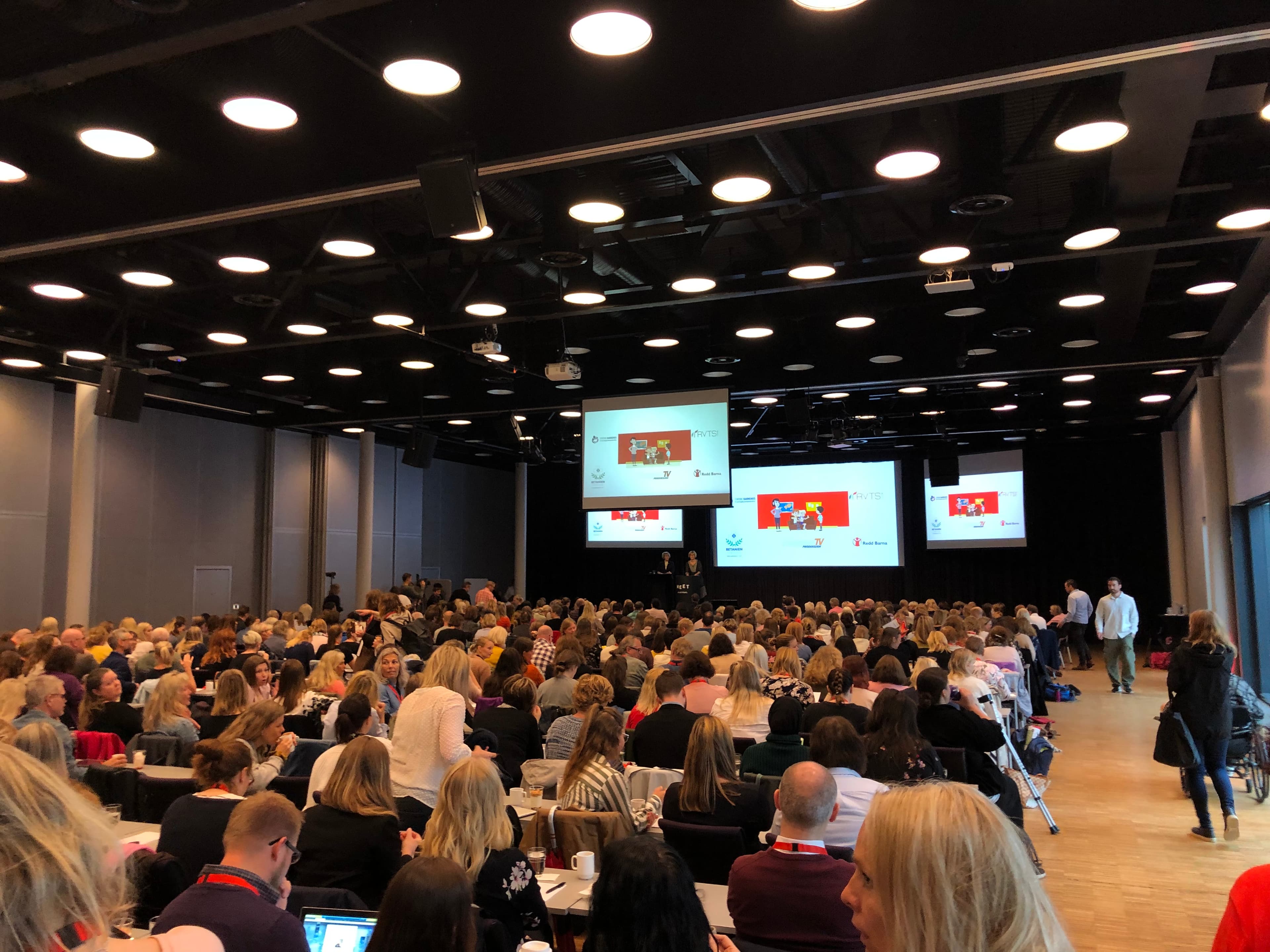 Et stort konferanserom fylt med mennesker som sitter og ser på presentasjoner som vises på flere skjermer foran i rommet. Atmosfæren virker travel og oppmerksom.