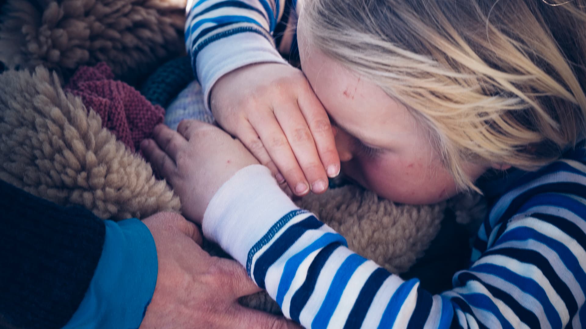Et barn med blondt hår, iført en blå- og hvitstripet skjorte, ligger med hendene over deler av ansiktet og hviler på et loddent teppe. En voksen hånd berører forsiktig barnets arm og gir trøst.