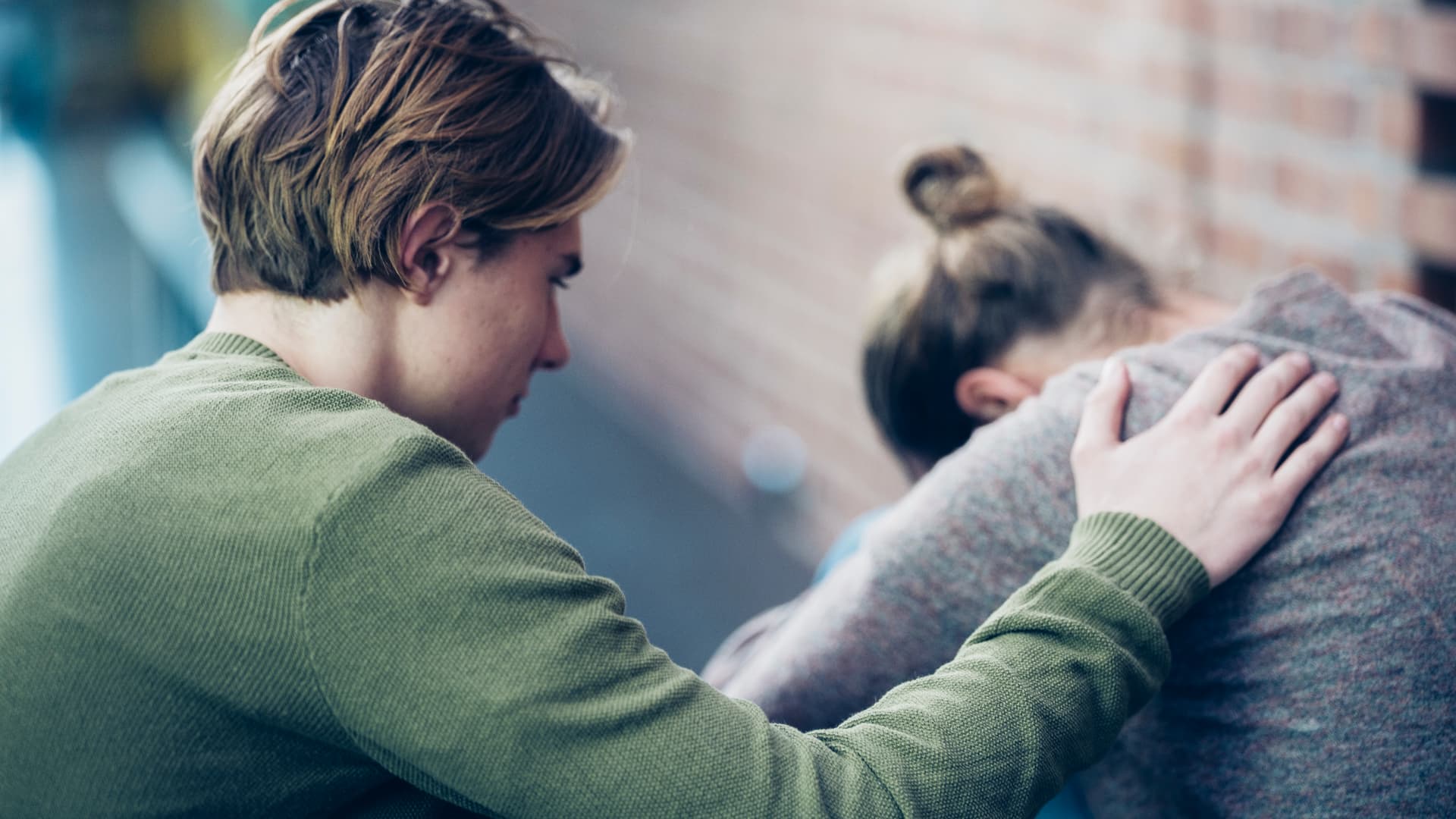 En person trøster en annen ved å legge en hånd på ryggen hans eller hennes. Den som blir trøstet, lener seg fremover med hodet bøyd, mens det i bakgrunnen ses en murvegg.