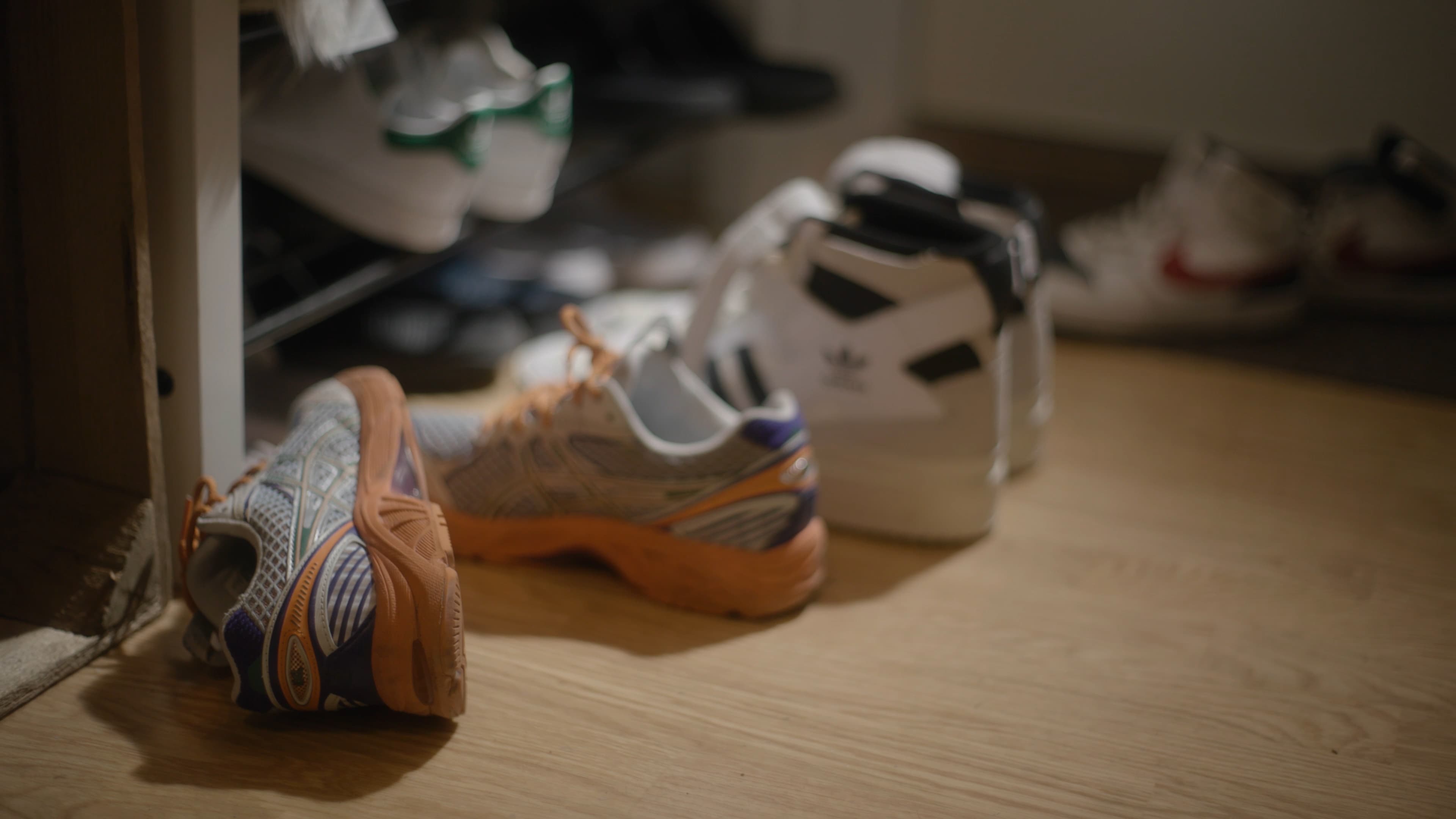 Et par slitte joggesko og et par hvite joggesko med svarte striper står på et tregulv, og flere sko er synlige på et stativ og i den uskarpe bakgrunnen.