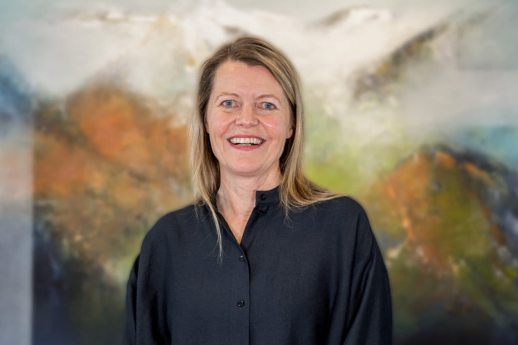 En kvinne med blondt hår og mørk skjorte med knapper smiler til kameraet. Bakgrunnen er et uskarpt, fargerikt, abstrakt maleri med jordfarger og innslag av hvitt og blått.