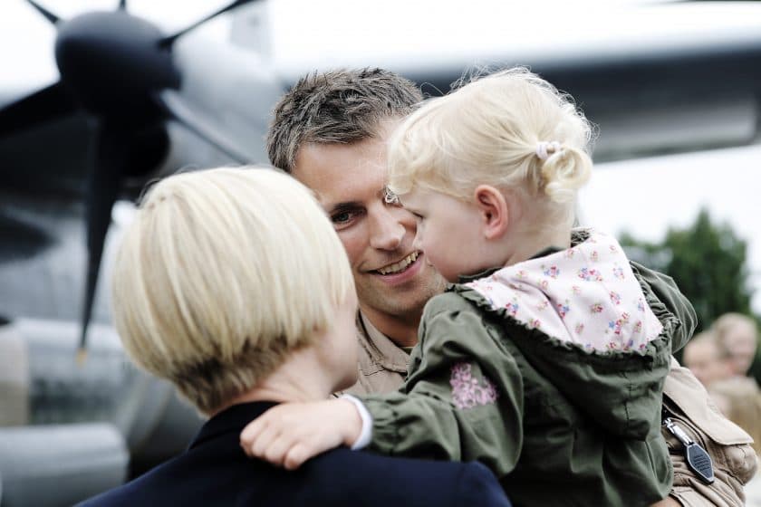 En smilende mann i militæruniform hilser på en kvinne og en ung, blond jente som blir holdt fast foran et stort fly. Scenen ser ut til å fange en varm gjenforening.