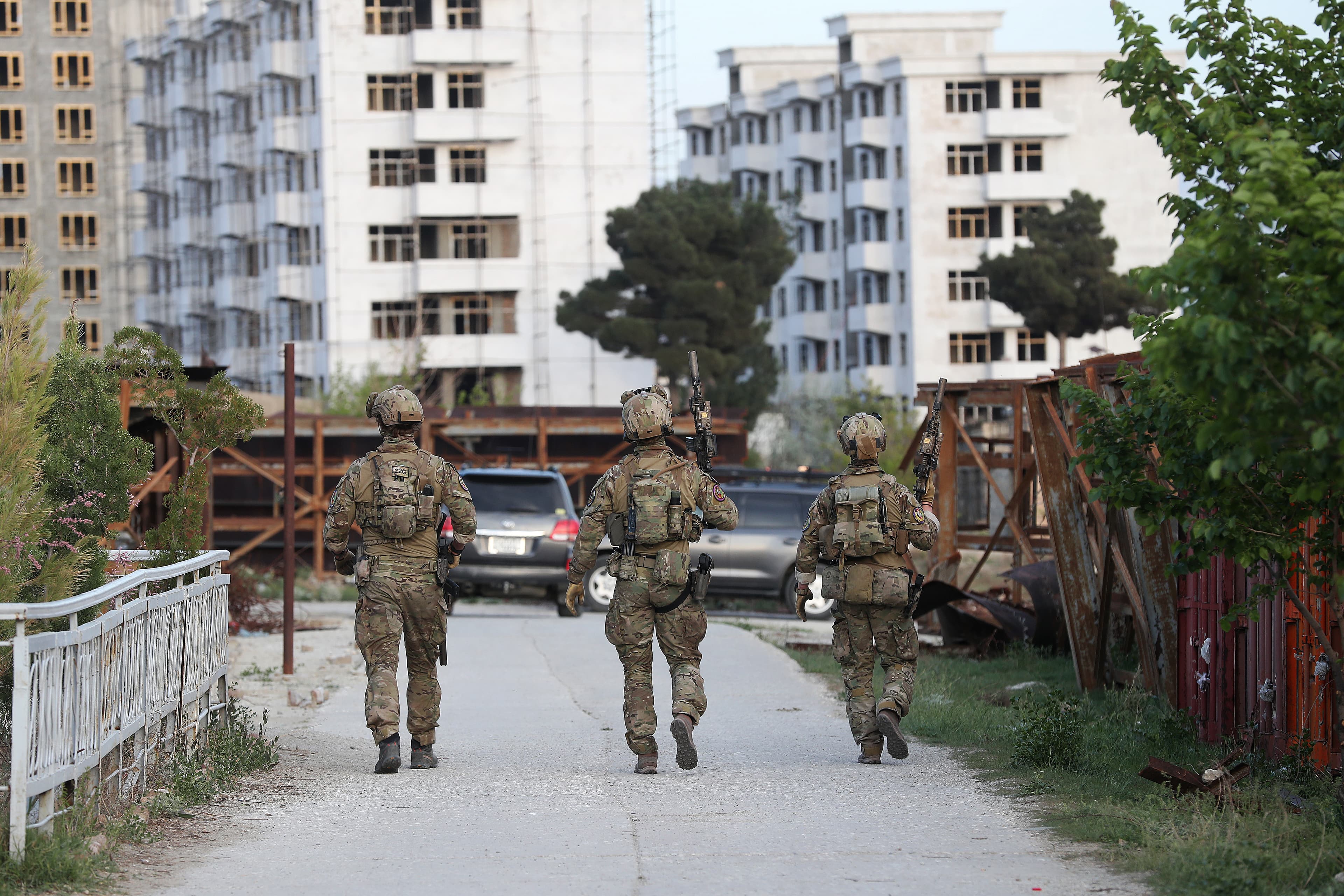 Tre soldater i kamputstyr går nedover en vei mot uferdige eller ødelagte boligblokker, med parkerte biler og sparsomt med trær på hver side, i et urbant område.