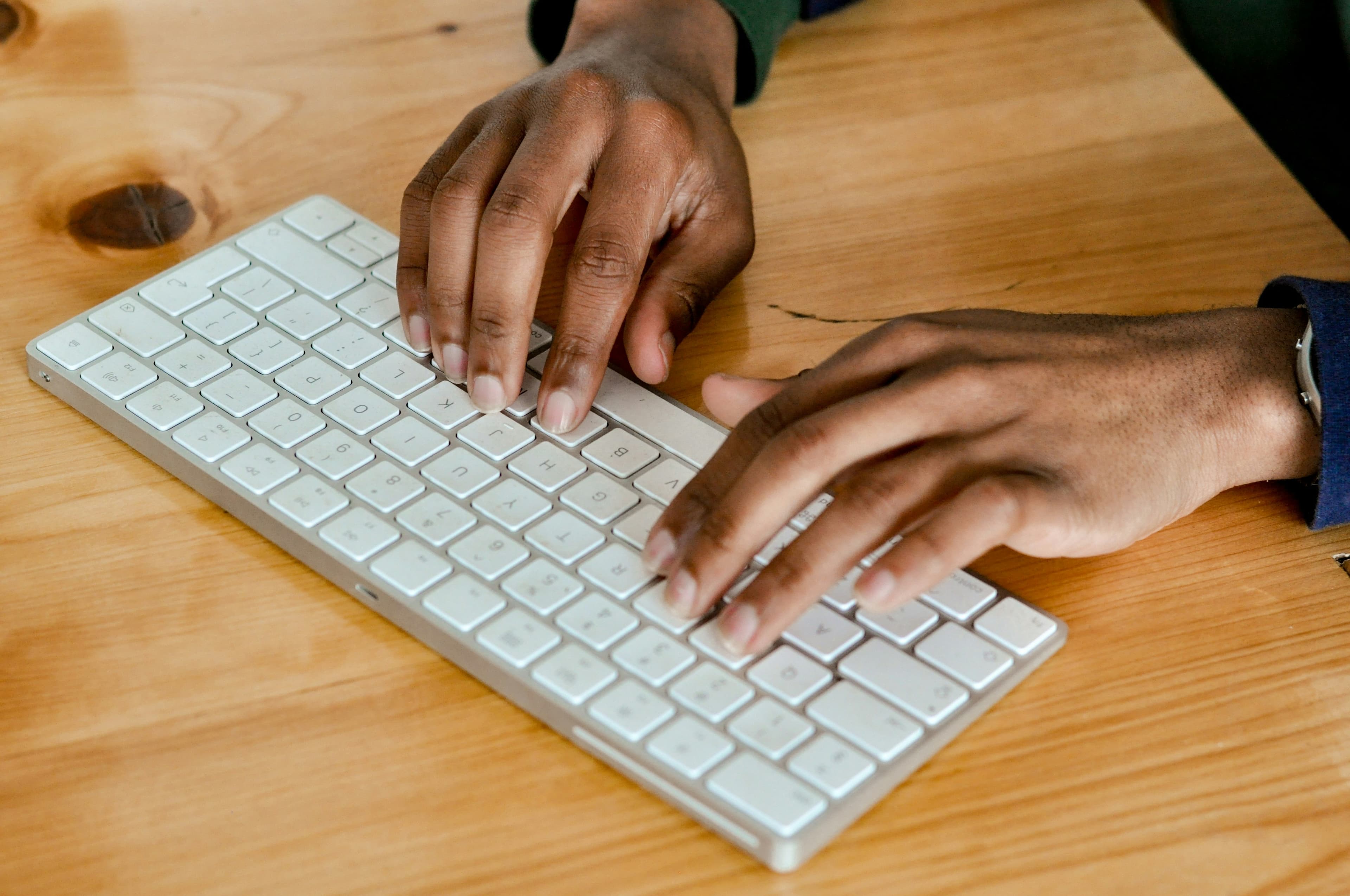 En person skriver på et slankt, hvitt, trådløst tastatur på et skrivebord av tre, med begge hender synlige.