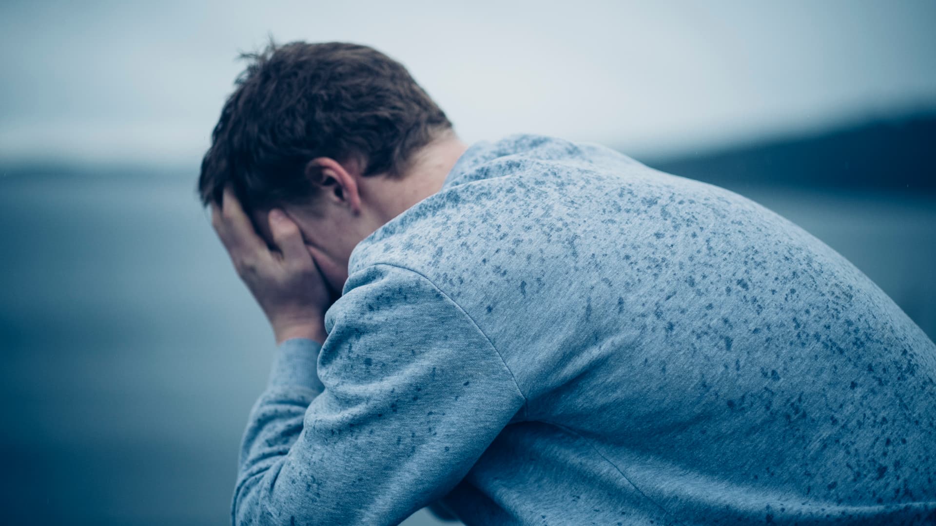 En person i grå genser sitter utendørs i regnet og dekker ansiktet med begge hender, og virker bedrøvet eller opprørt. Bakgrunnen er uskarp, med dempede blå og grå toner.