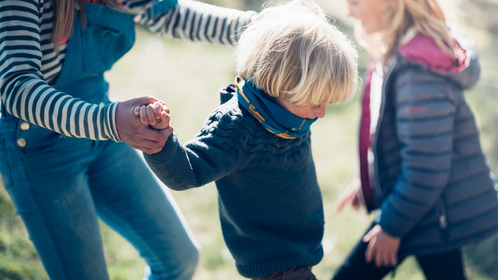 Et lite barn med blondt hår, iført genser og skjerf, holder en voksen i hånden for å få støtte mens hun går utendørs. Et annet barn i jakke står i nærheten. Scenen er sterkt opplyst og virker leken.