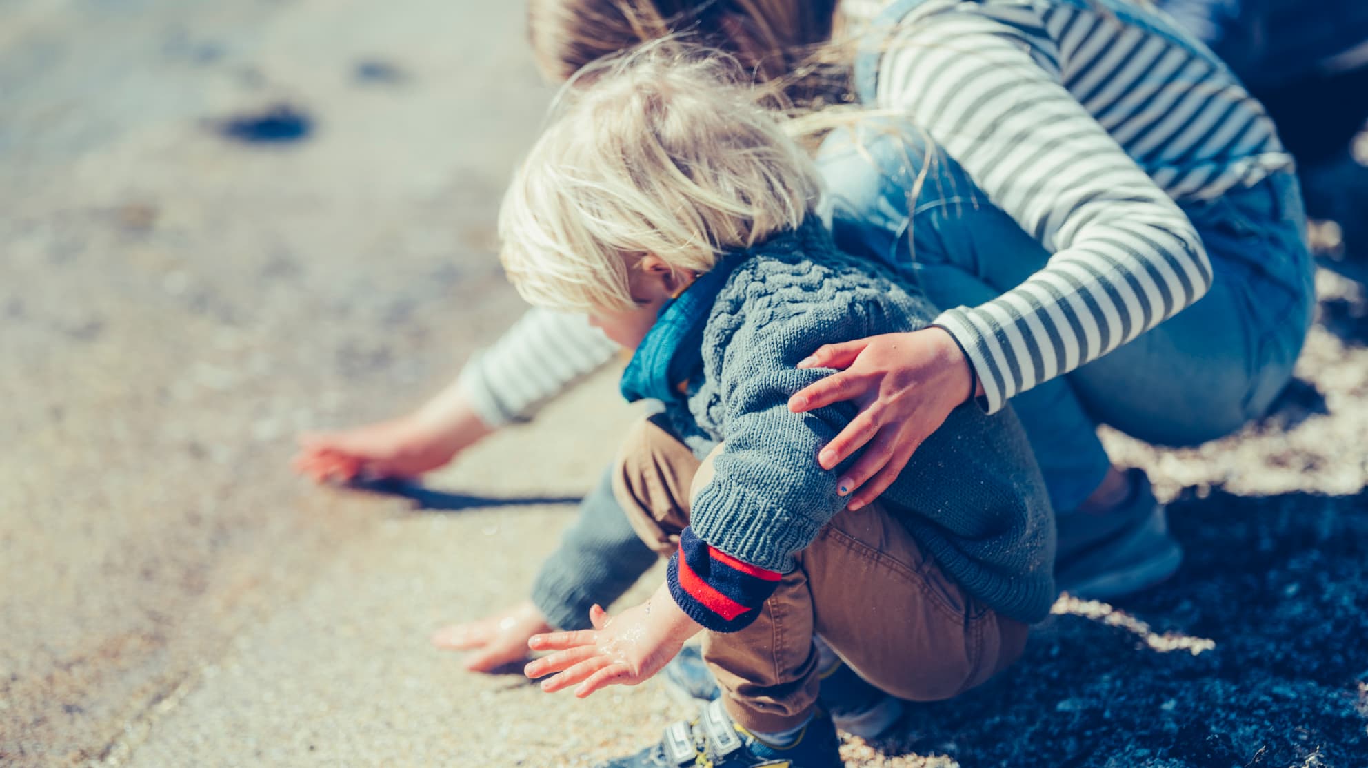 Et lite barn med blondt hår sitter på huk på en sandstrand nær vannkanten og strekker seg mot sanden, mens en voksen ved siden av forsiktig legger en hånd på barnets rygg. Begge er kledd i varme klær.