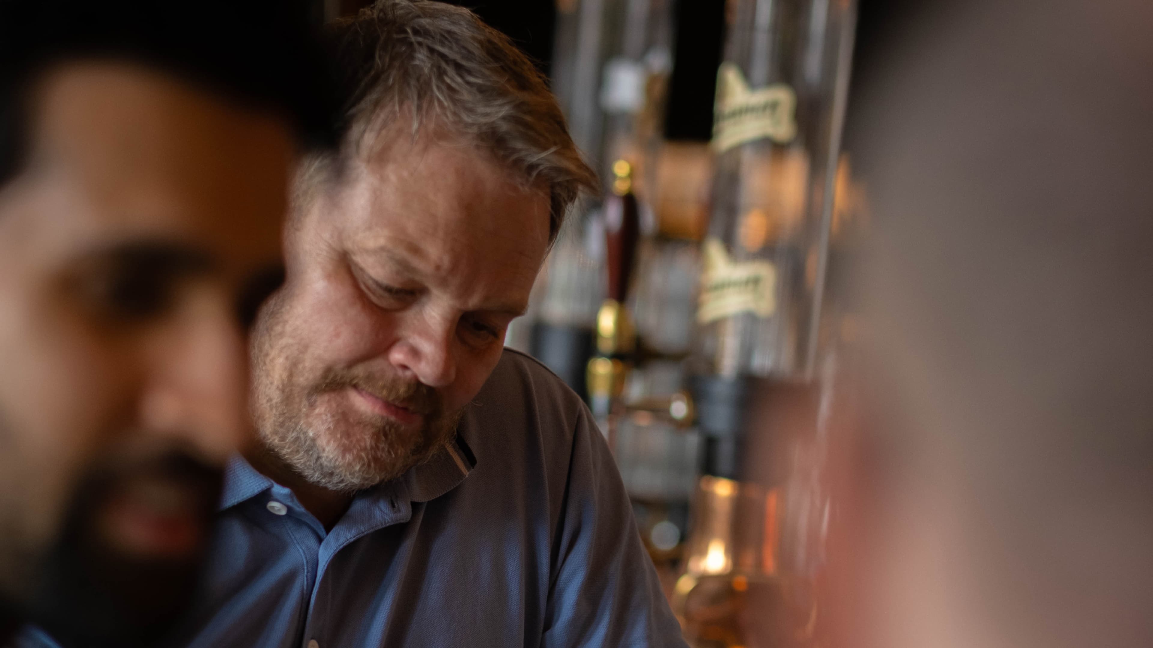 En middelaldrende mann med lyst hår og skjegg ser tankefullt ned i en svakt opplyst bar, med ølkraner og uskarpe menneskefigurer synlige både i forgrunnen og bakgrunnen.