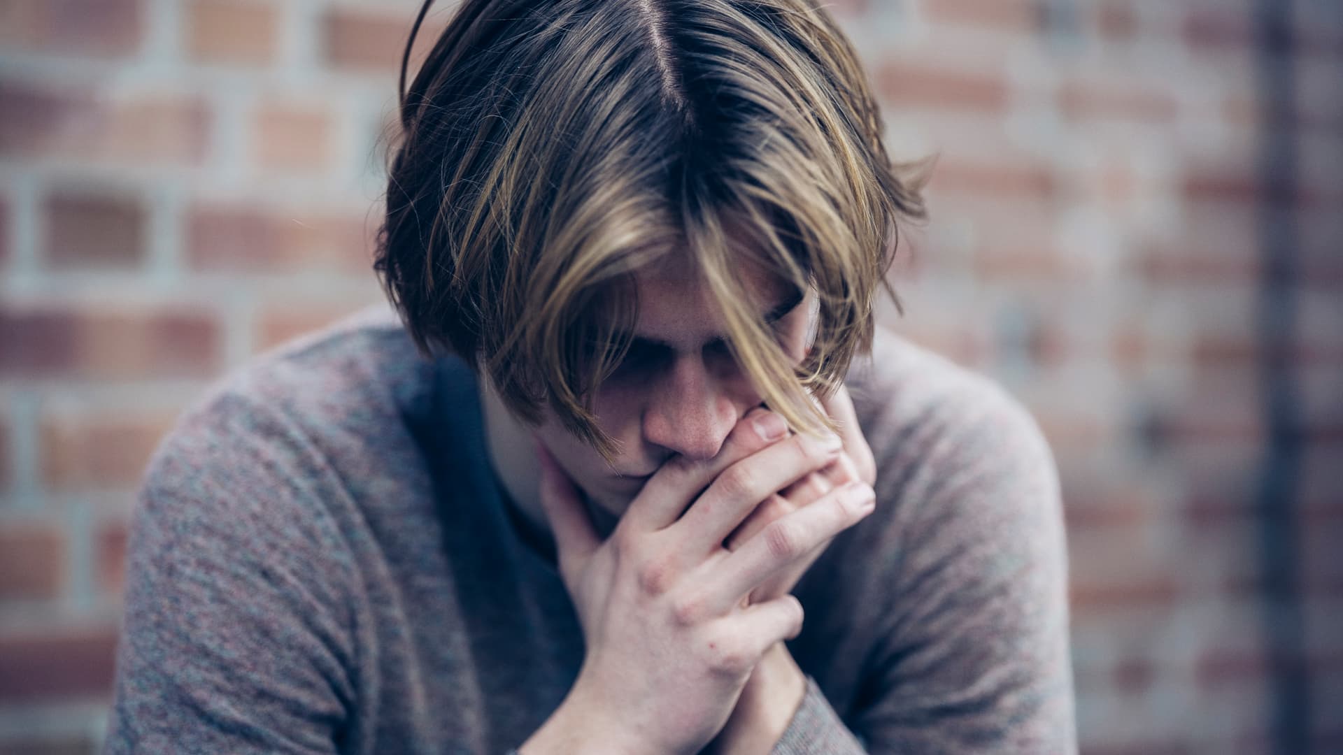 En person med blondt hår sitter med hodet ned og hendene for munnen, og ser ut til å være dypt i tanker eller stresset, mot en uskarp bakgrunn av en murvegg.