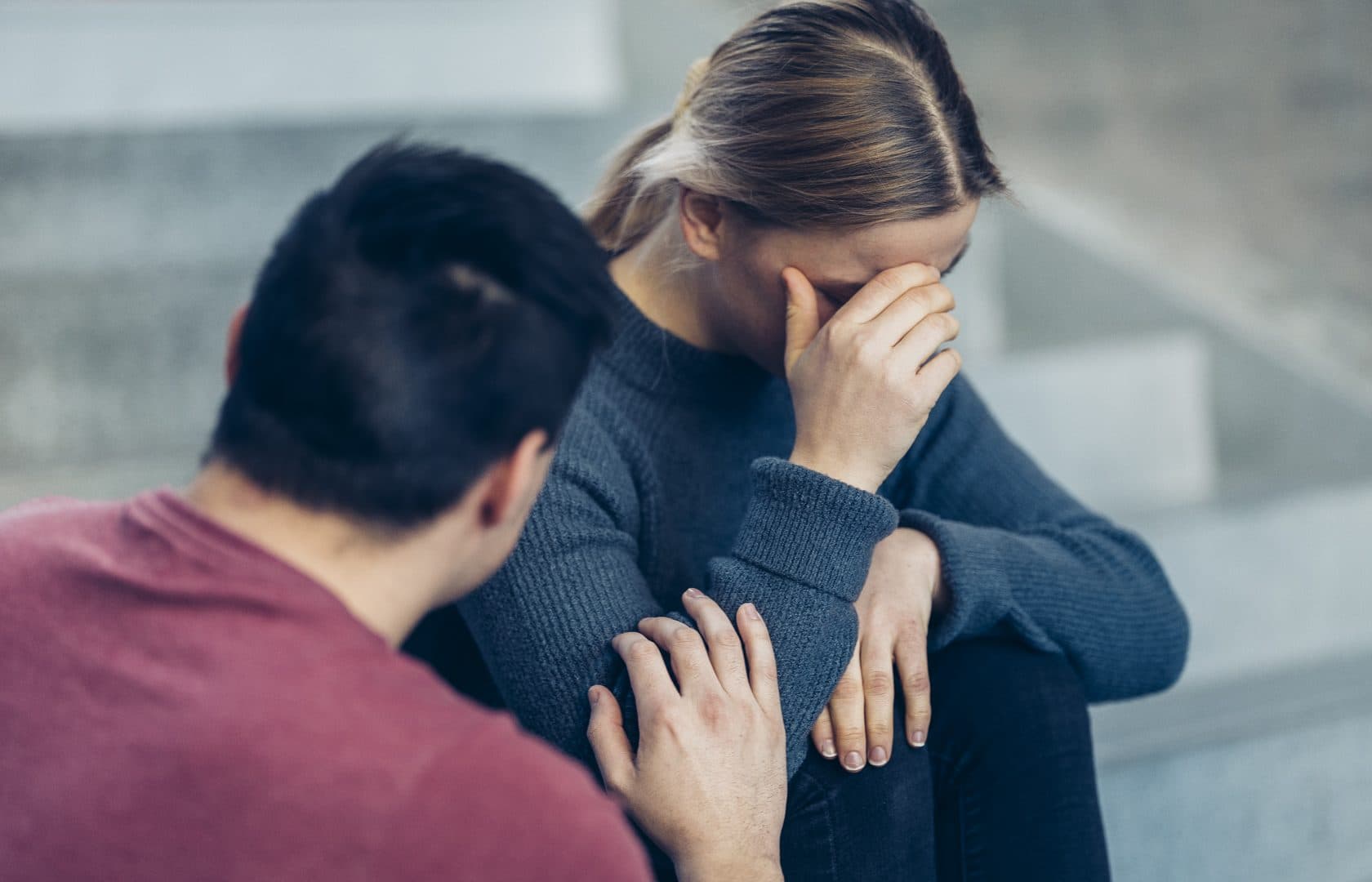 En kvinne sitter på en trapp og dekker ansiktet med hånden i nød. En mann ved siden av henne berører forsiktig armen hennes og tilbyr trøst og støtte.