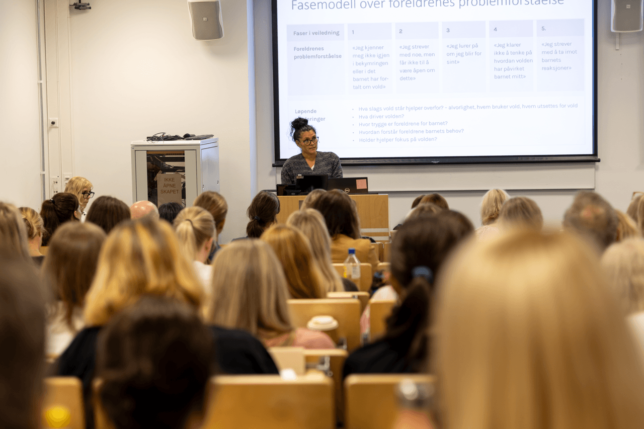 En kvinne står ved en talerstol og holder et foredrag for et klasserom fullt av sittende mennesker. Bak henne vises det på en stor skjerm et lysbilde med tekst på dansk om faser i foreldres problemsituasjoner.