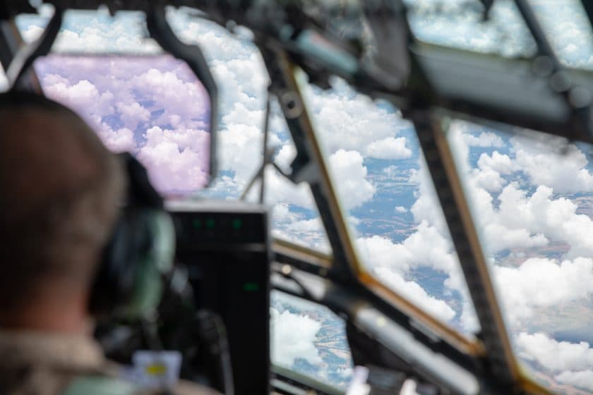 Utsikt fra innsiden av en flycockpit med en uskarp pilot i forgrunnen og en klar himmel fylt med hvite skyer gjennom vinduene.
