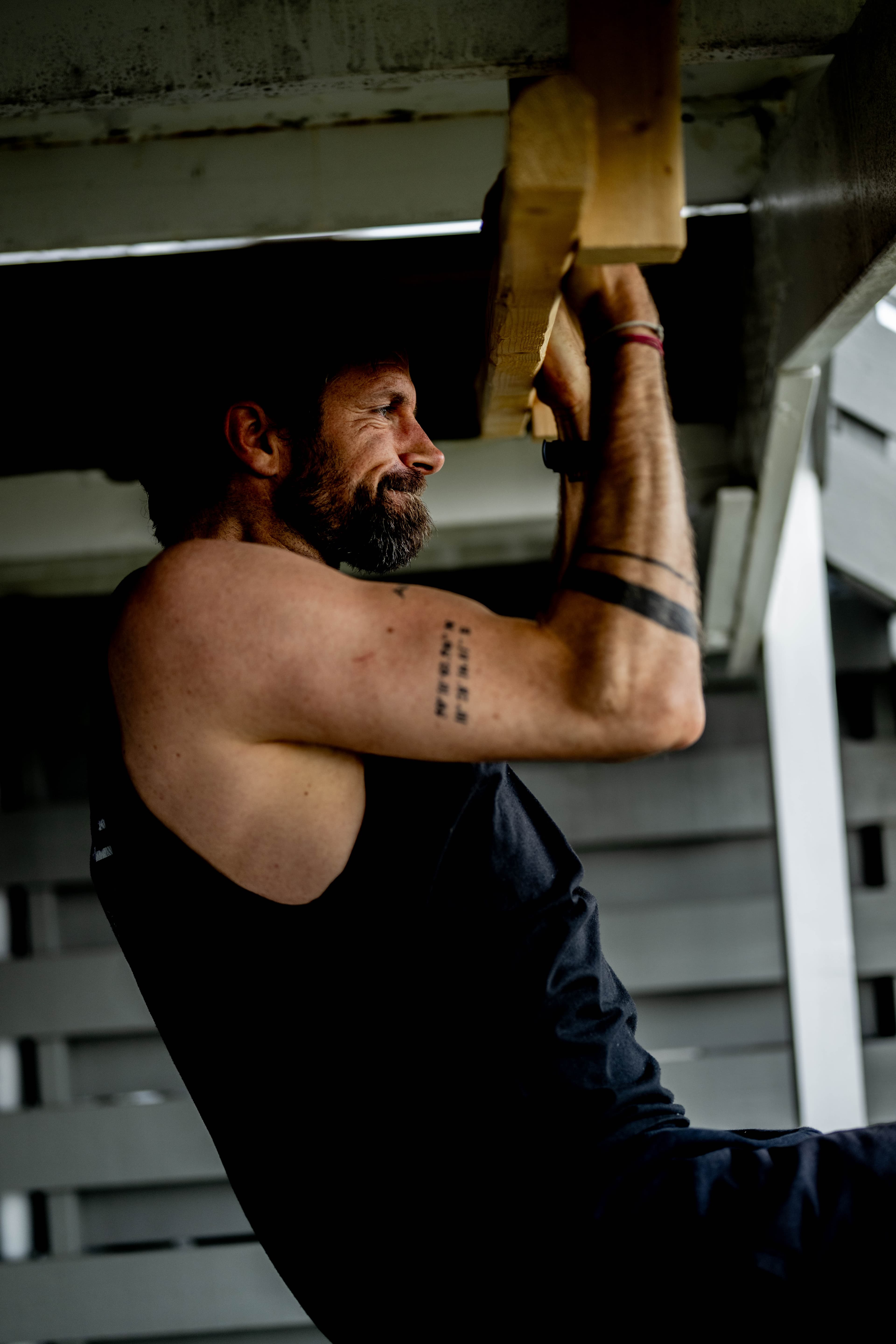 En mann med skjegg og tatoveringer på armen gjør en pull-up på en trebjelke utendørs, iført en svart tank top.