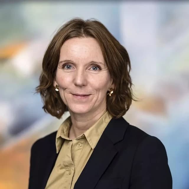 En kvinne med lysebrunt hår, kledd i en beige skjorte og en mørk blazer, smiler svakt foran en uskarp, fargerik bakgrunn.
