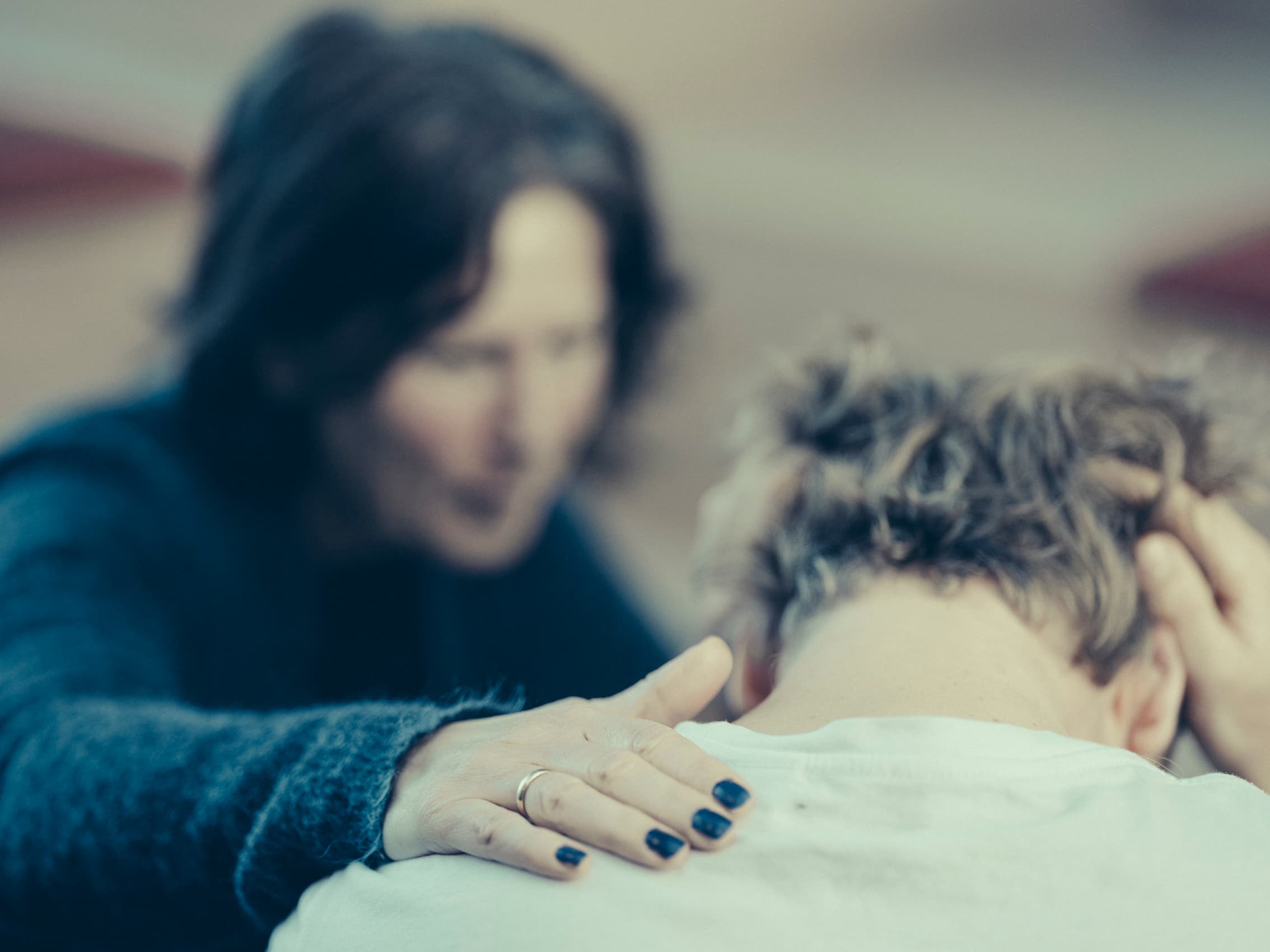 En mørkhåret person legger forsiktig en trøstende hånd på skulderen til en annen person som sitter med hodet ned og hendene på hodet, noe som tyder på emosjonell støtte eller trøst.