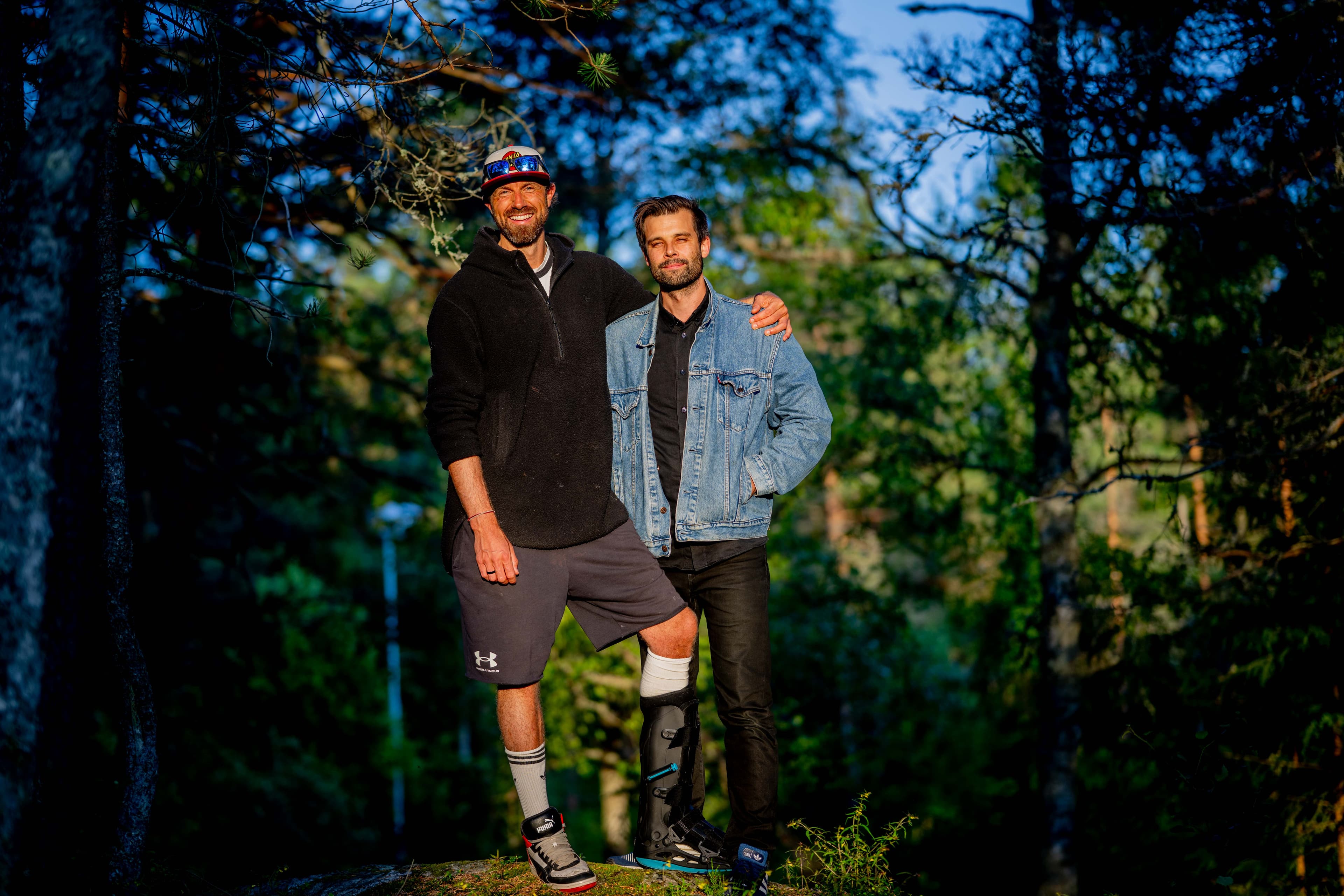 To menn står sammen i en solopplyst skog og smiler mot kameraet. Den ene mannen har benprotese og er iført svart jakke, shorts og lue, mens den andre er iført dongerijakke og svarte bukser. De er omgitt av trær.