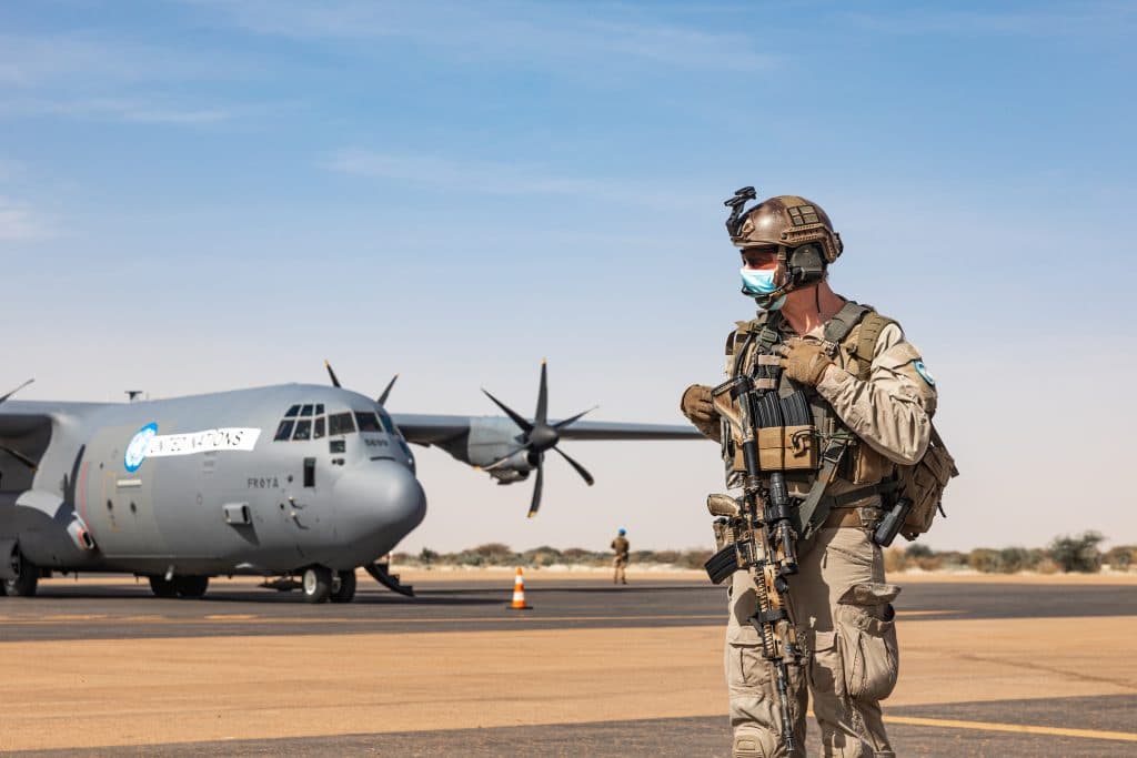 En soldat iført taktisk utstyr og ansiktsmaske holder vakt på en flystripe med et gevær i hånden, mens et militært transportfly står parkert i bakgrunnen under en klar himmel.