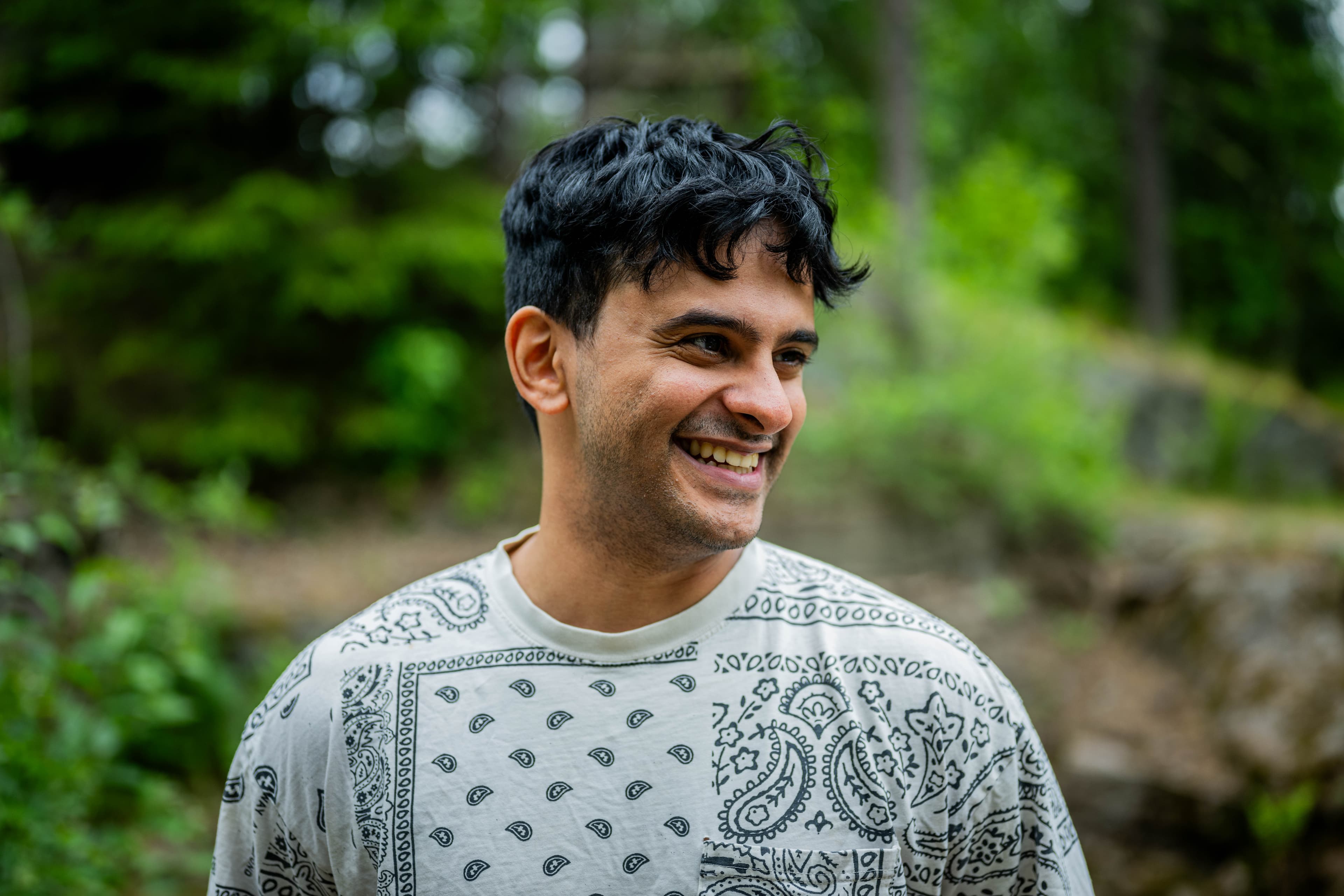 En mann med mørkt hår smiler mens han står utendørs i et grønt, skogkledd område. Han har på seg en lys skjorte med svart paisleymønster. Bakgrunnen er uskarp med trær og løvverk.