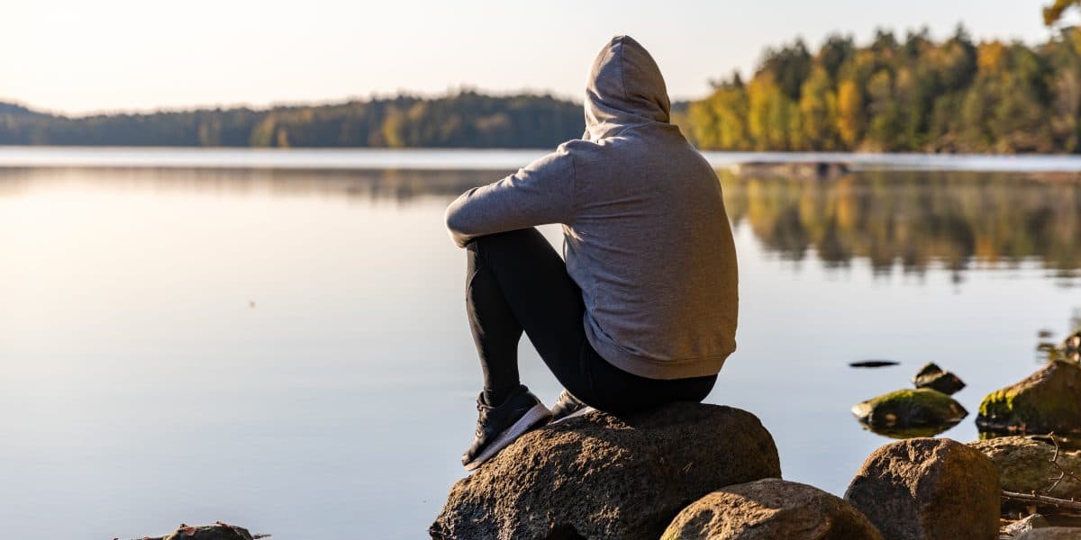 En person i grå hettegenser sitter på en stein ved en rolig innsjø, vendt mot vannet og trærne i det fjerne, og ser ut til å reflektere eller slappe av i de fredelige, naturlige omgivelsene.