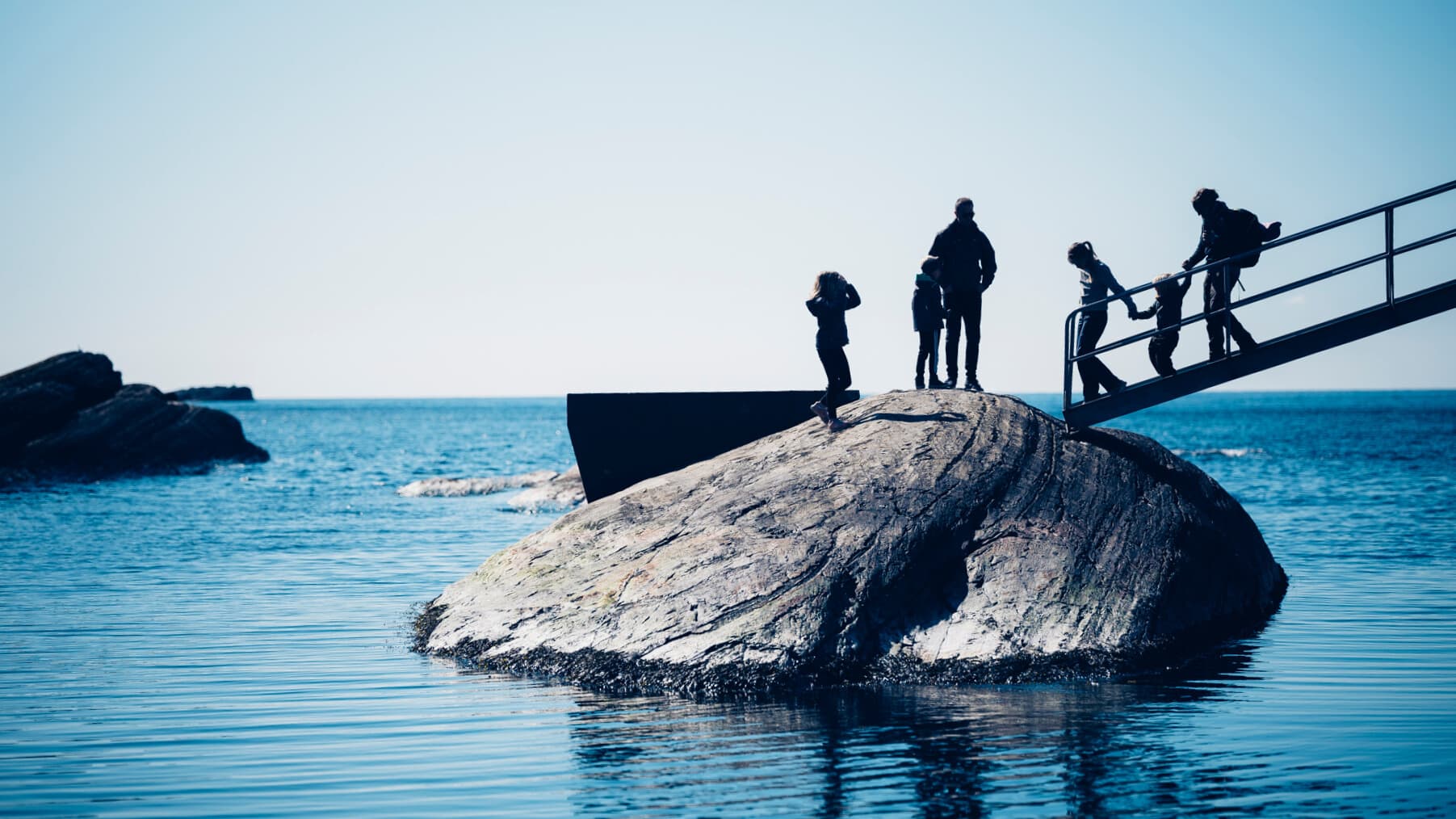 En gruppe mennesker står og går på en stor stein i havet, og noen går ned en metalltrapp ned på steinen. Himmelen er klar, og vannet er rolig og blått.