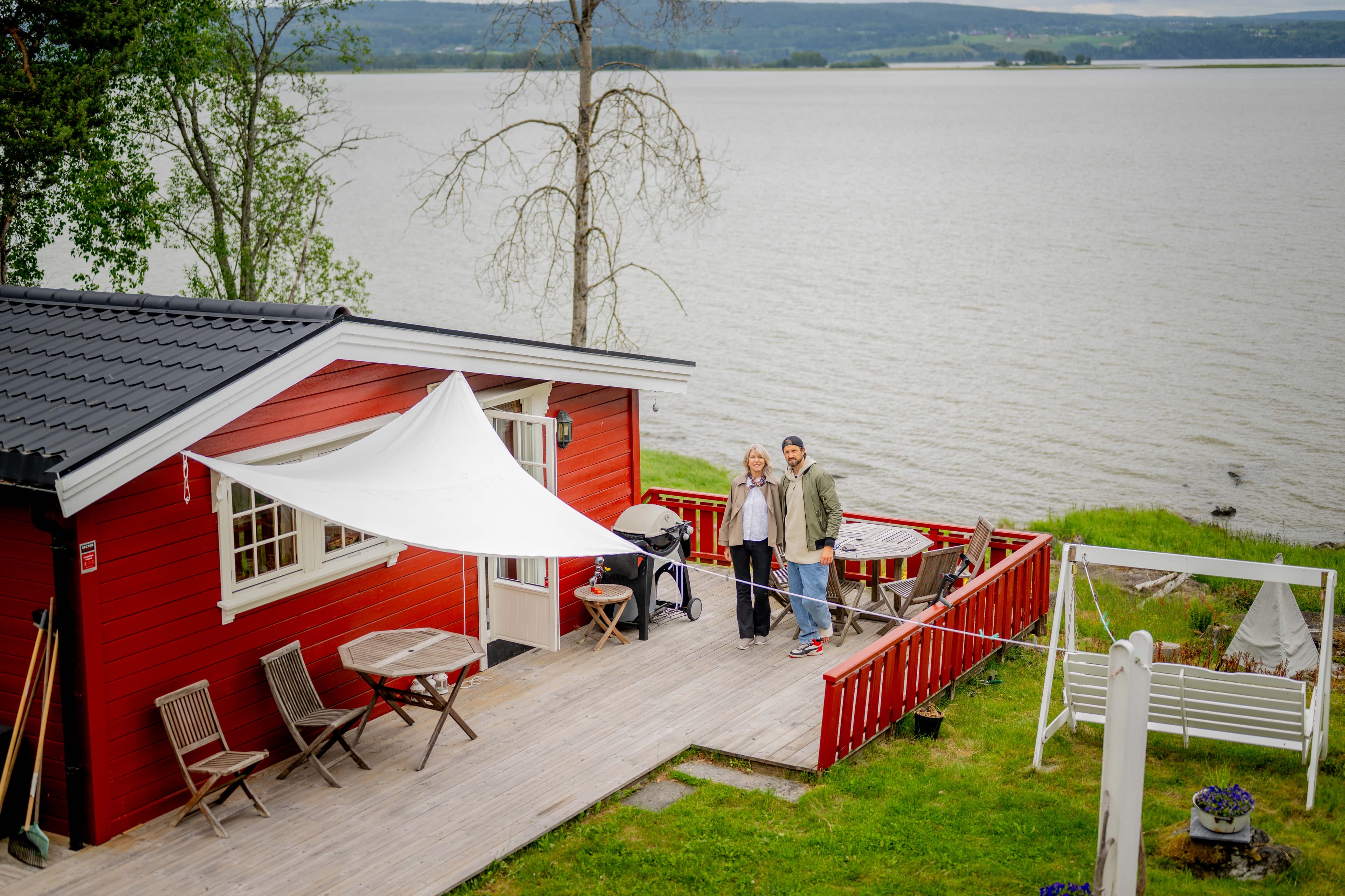 Et par står på tredekket til en rød hytte ved innsjøen, med utemøbler, grill og et hvitt tak. Scenen har utsikt over en rolig innsjø og et grønt landskap under en overskyet himmel.