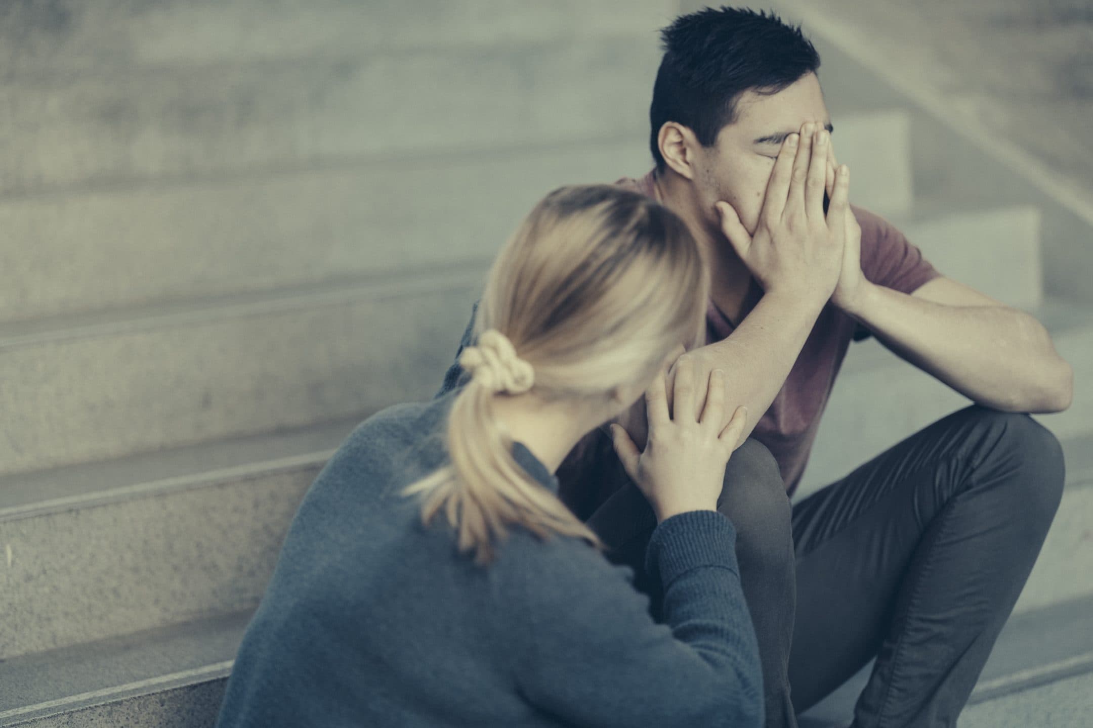 En kvinne sitter ved siden av en mann på en trapp og holder ham i hånden. Mannen dekker ansiktet med begge hender og virker fortvilet, mens kvinnen ser ut til å trøste ham.