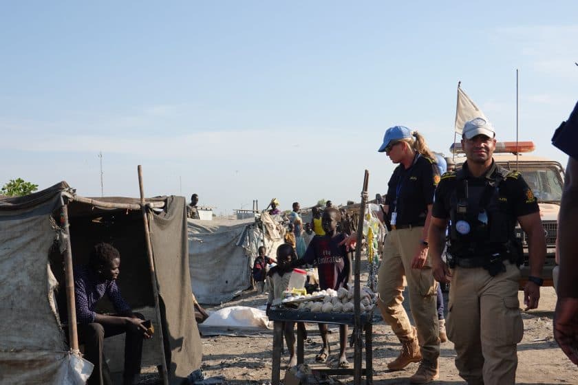 To hjelpearbeidere går gjennom en flyktningleir og passerer provisoriske telt. Barn står i nærheten, ett av dem selger egg, en annen sitter inne i et telt. Stemningen virker varm og solrik.