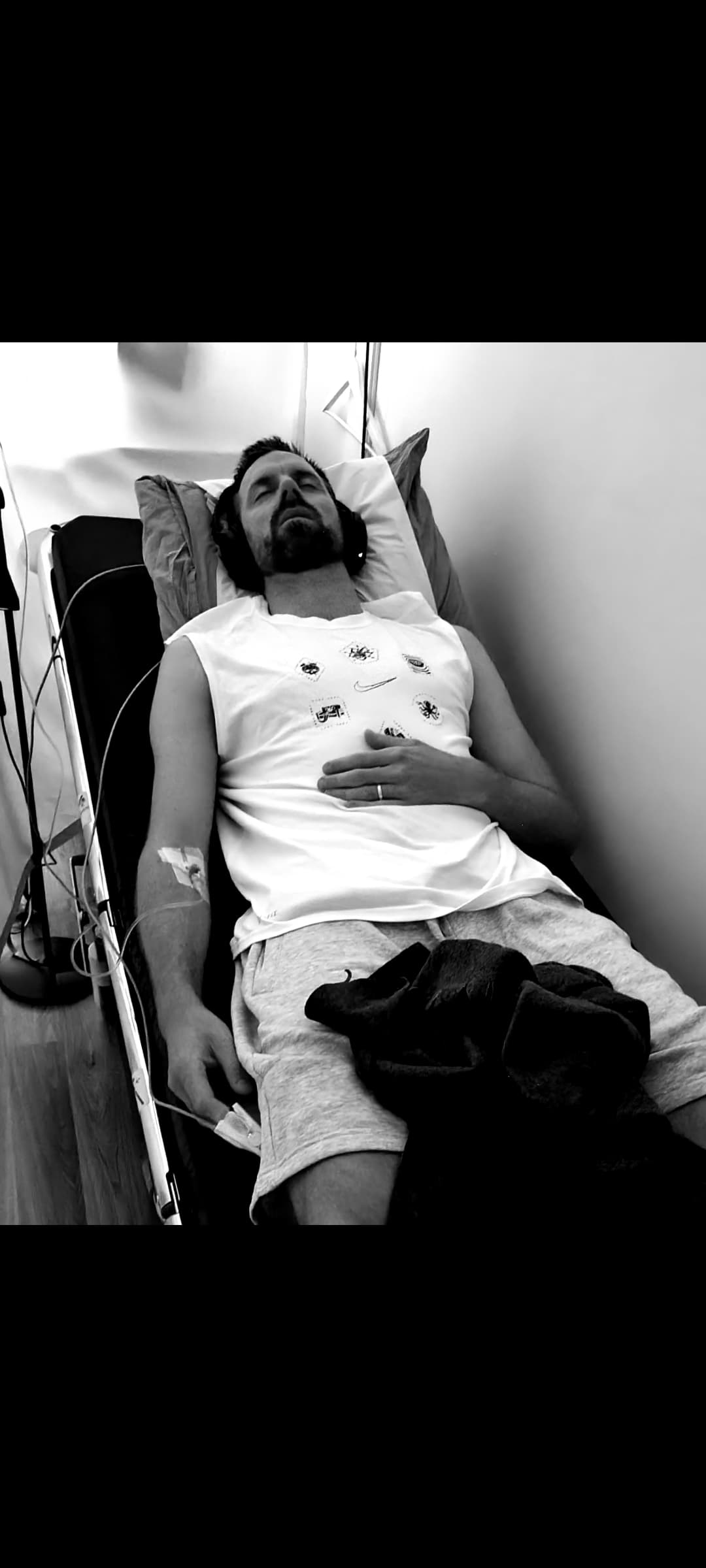 En mann ligger på en sykehusseng med medisinske monitorer festet til brystet og et intravenøst system i armen. Han ser ut til å hvile eller være bevisstløs, iført T-skjorte og shorts. Bildet er i svart-hvitt.