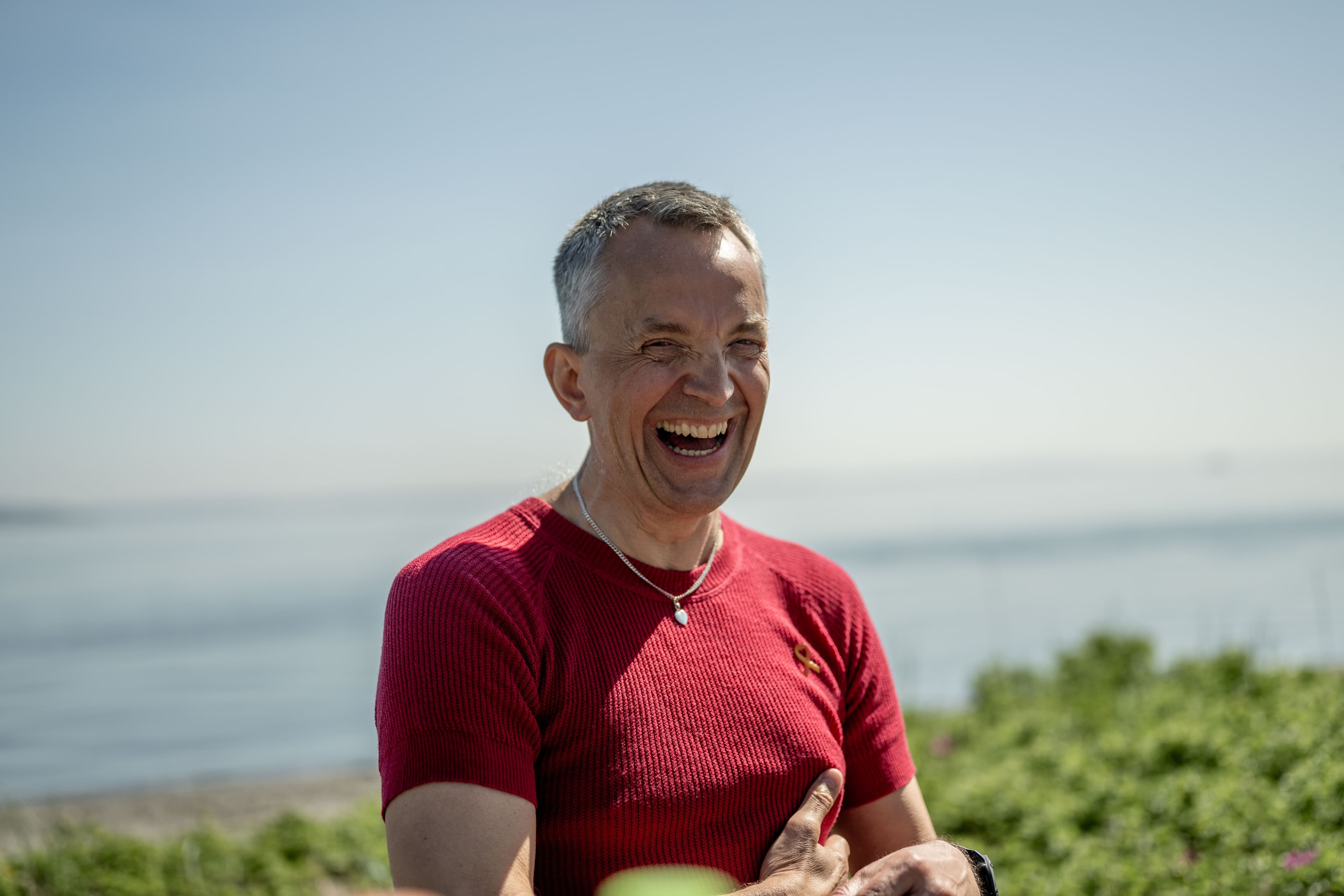 En mann i rød skjorte og halskjede ler fornøyd utendørs, med grønne planter og et stille vann i bakgrunnen under en klarblå himmel.