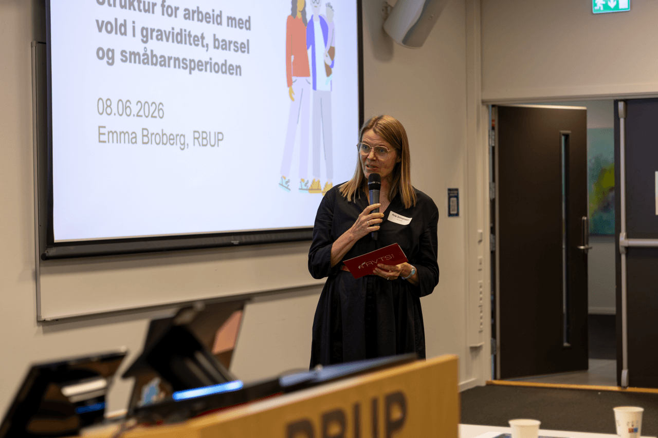 En kvinne med en mikrofon i hånden står foran en skjerm som viser en presentasjon med tittelen «Struktur for arbeid med vold i graviditet, barsel og småbarnsperioden», med navnet Emma Broberg, RBUP, og datoen 08.06.2026.