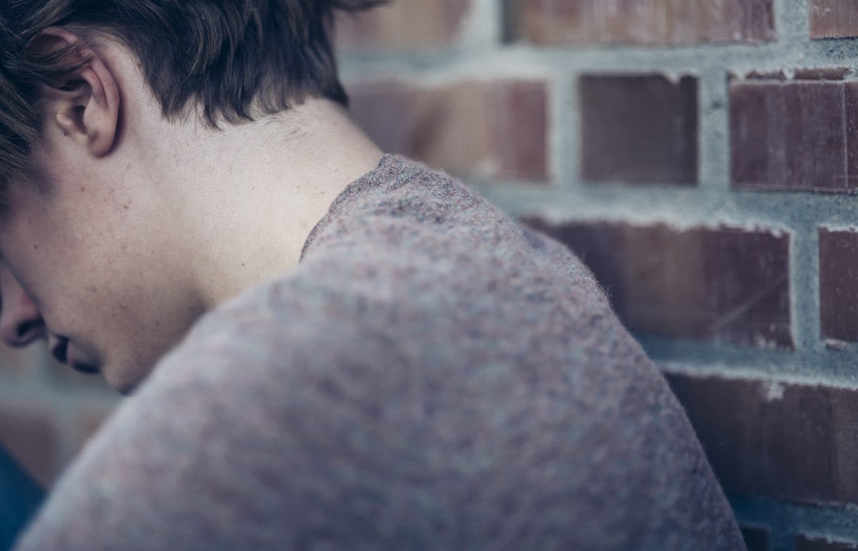 En person med kort, brunt hår og grå genser sitter med hodet ned og ryggen vendt mot kameraet mot en murvegg, noe som tyder på en stemning av tristhet eller kontemplasjon.