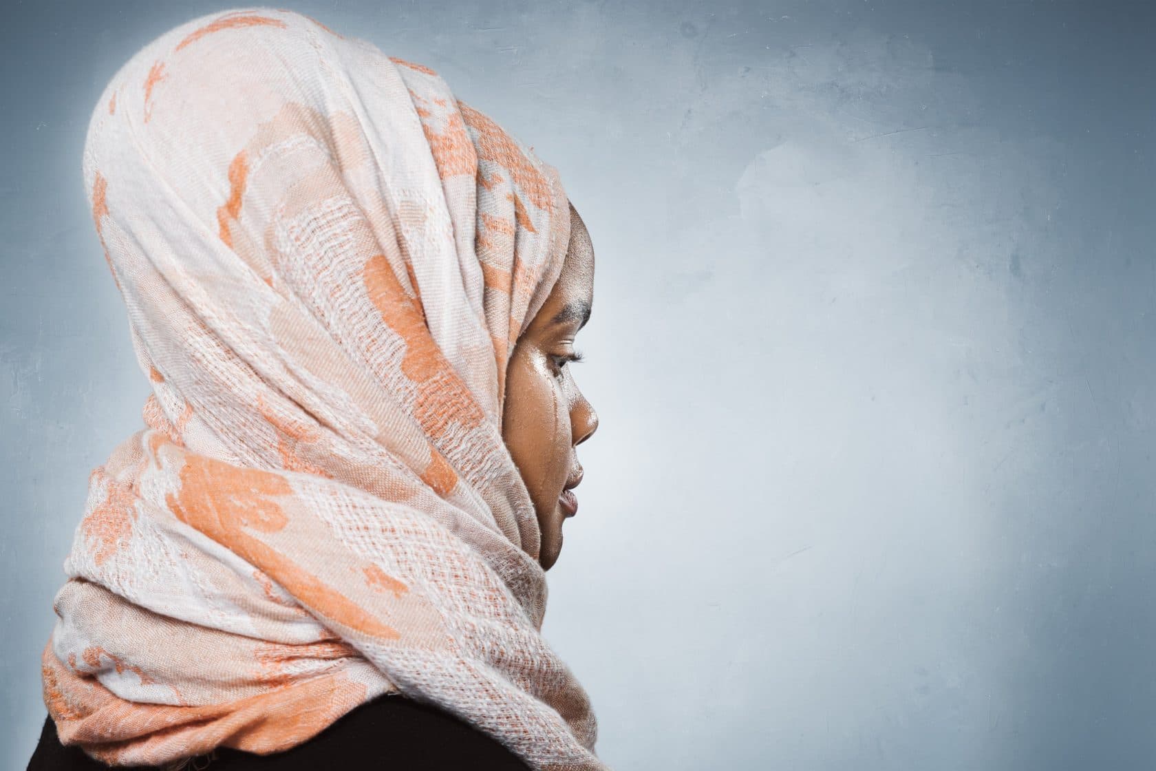 En ung kvinne iført en mønstret, lys hijab med oransje aksenter er avbildet i profil mot en ensfarget, lysegrå bakgrunn. Hun virker ettertenksom og har et rolig uttrykk.