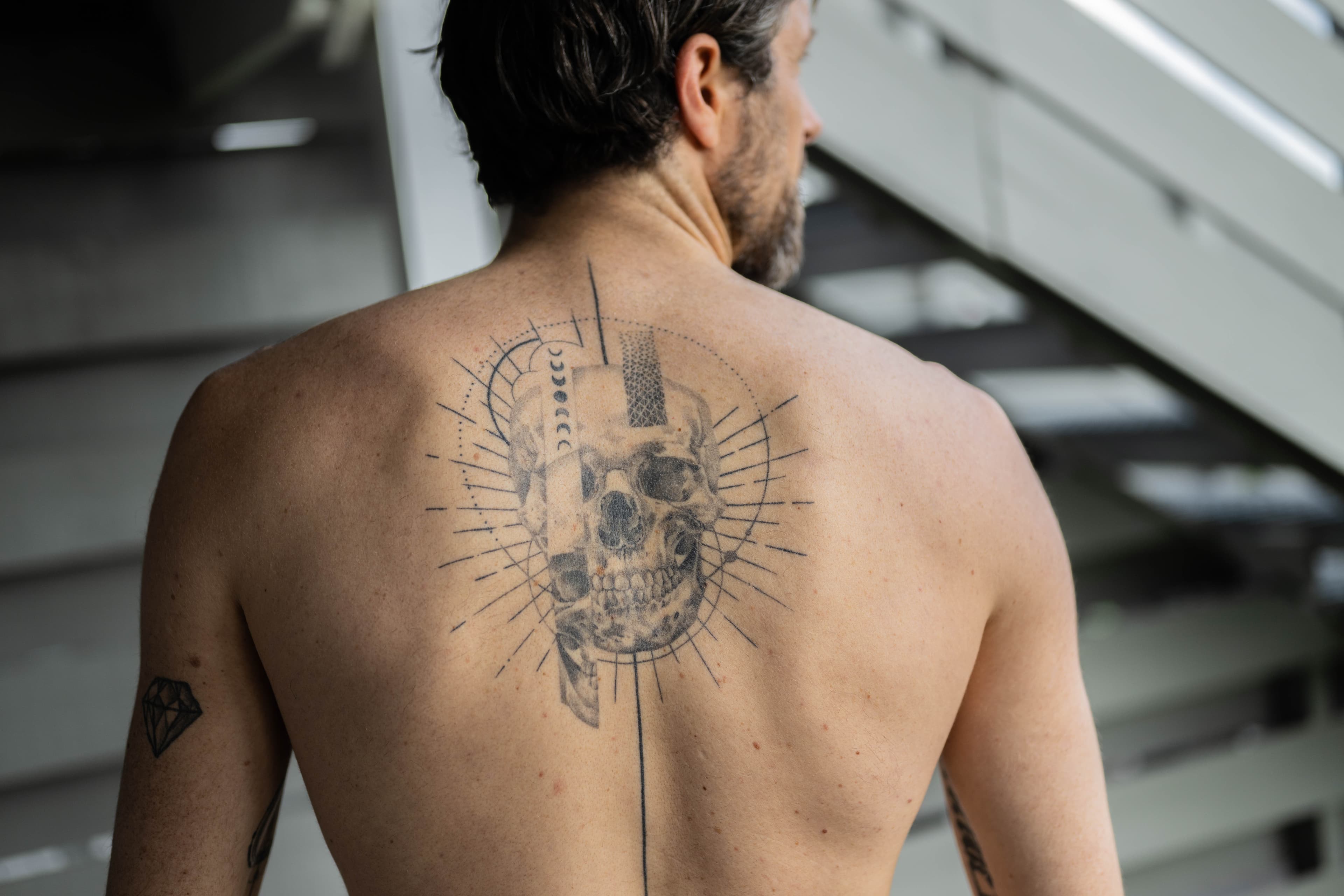 En mann med kort, mørkt hår viser frem sin nakne rygg, med en stor, svart tatovering av en geometrisk hodeskalle med radiale linjer og abstrakte elementer. Han står i nærheten av en trapp i et lyst innemiljø.