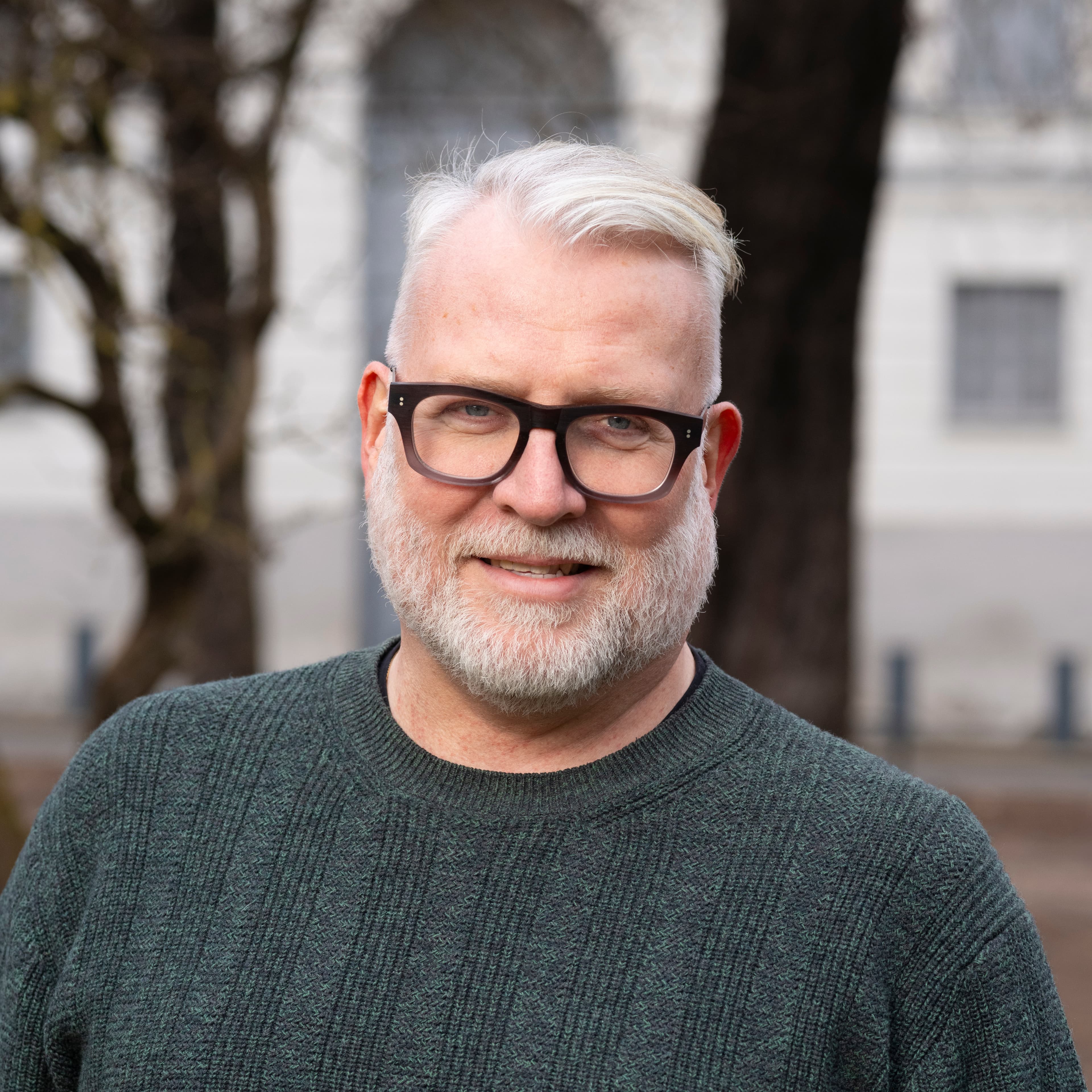 En middelaldrende mann med kort, hvitt hår, skjegg og briller smiler utendørs. Han har på seg en mørkegrønn genser, og trær og en lys bygning er uskarpe i bakgrunnen.