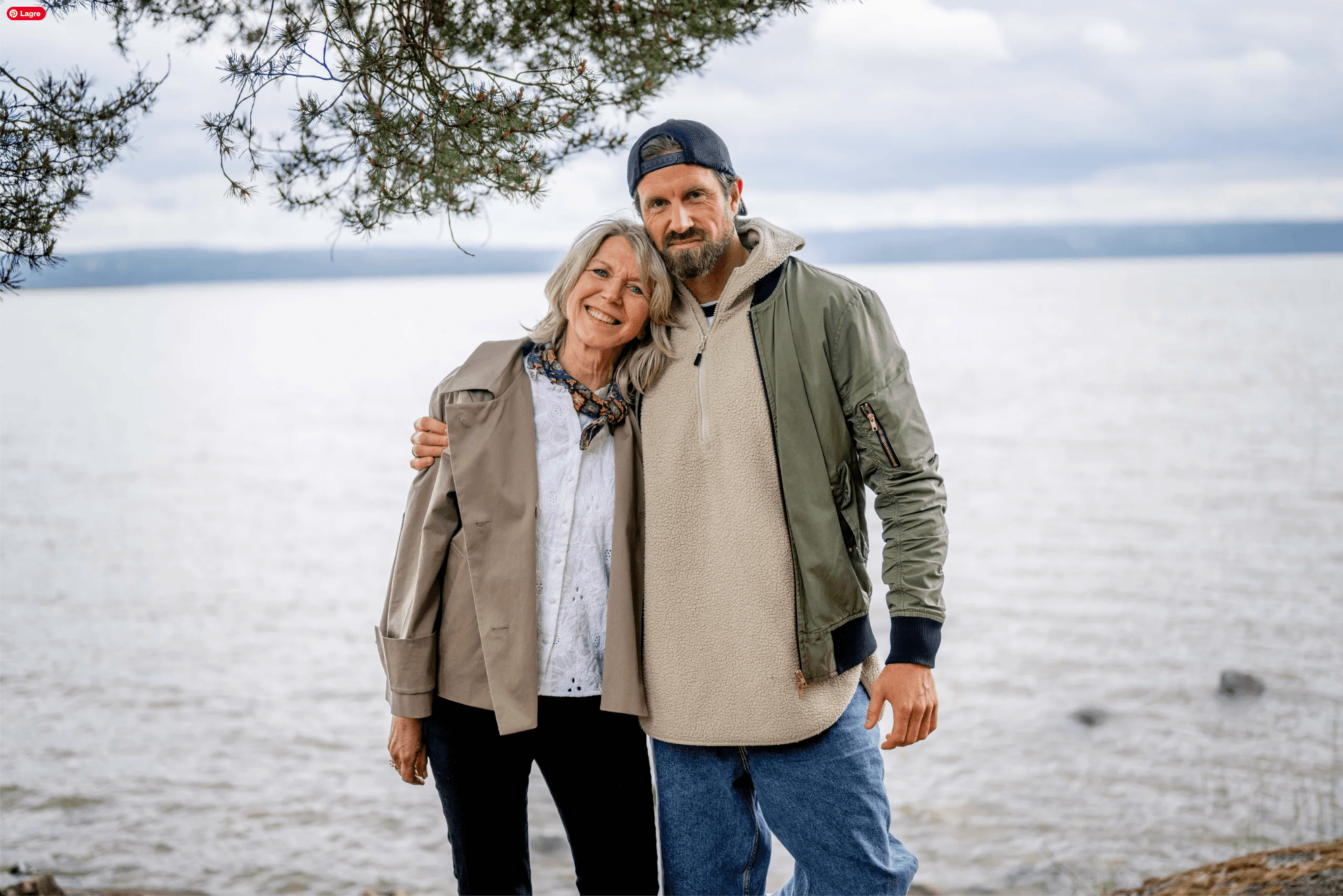 En eldre kvinne og en yngre mann står tett sammen, smilende, med armen hans rundt skuldrene hennes. De er utendørs ved en innsjø, med trær og overskyet himmel i bakgrunnen.