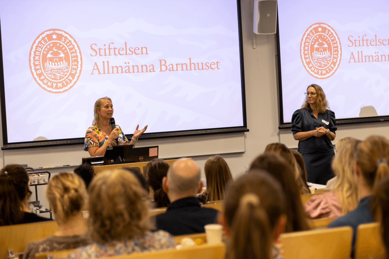 To kvinner står foran i en forelesningssal og snakker til publikum. På et presentasjonsbilde bak dem vises logoen og teksten «Stiftelsen Allmänna Barnhuset». Publikum lytter oppmerksomt.