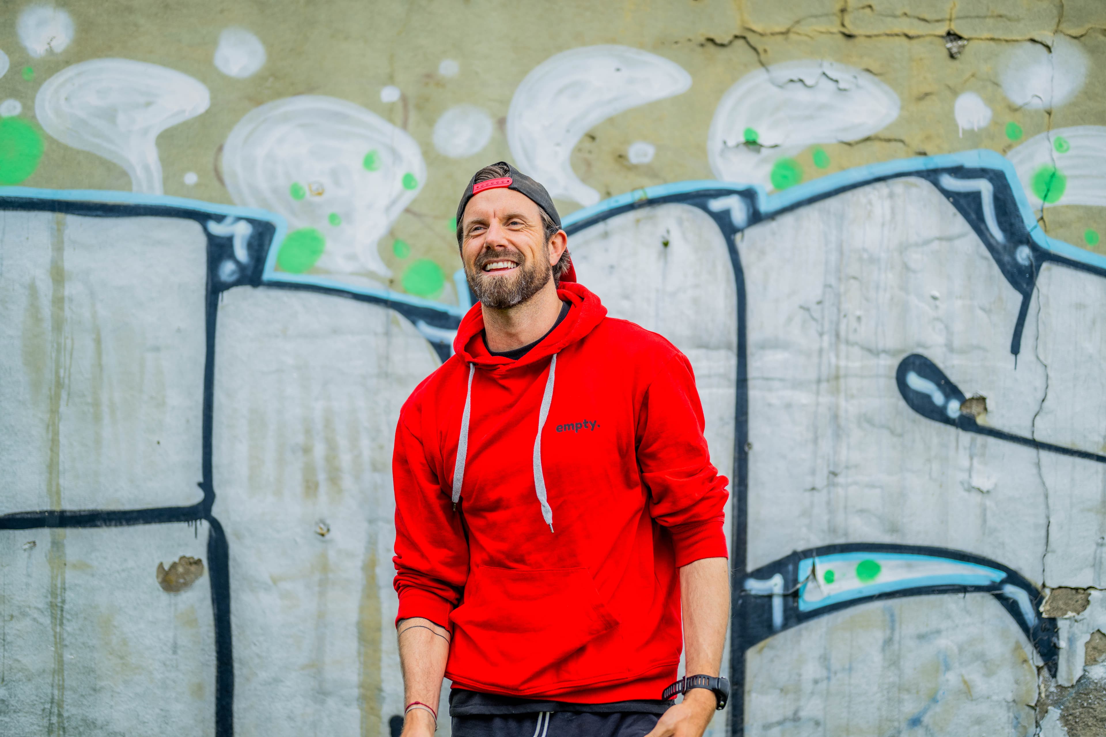 En mann i rød hettegenser og baklengs caps smiler mens han står foran en graffitikledd vegg. Bakgrunnen er preget av abstrakte former og dempede farger.