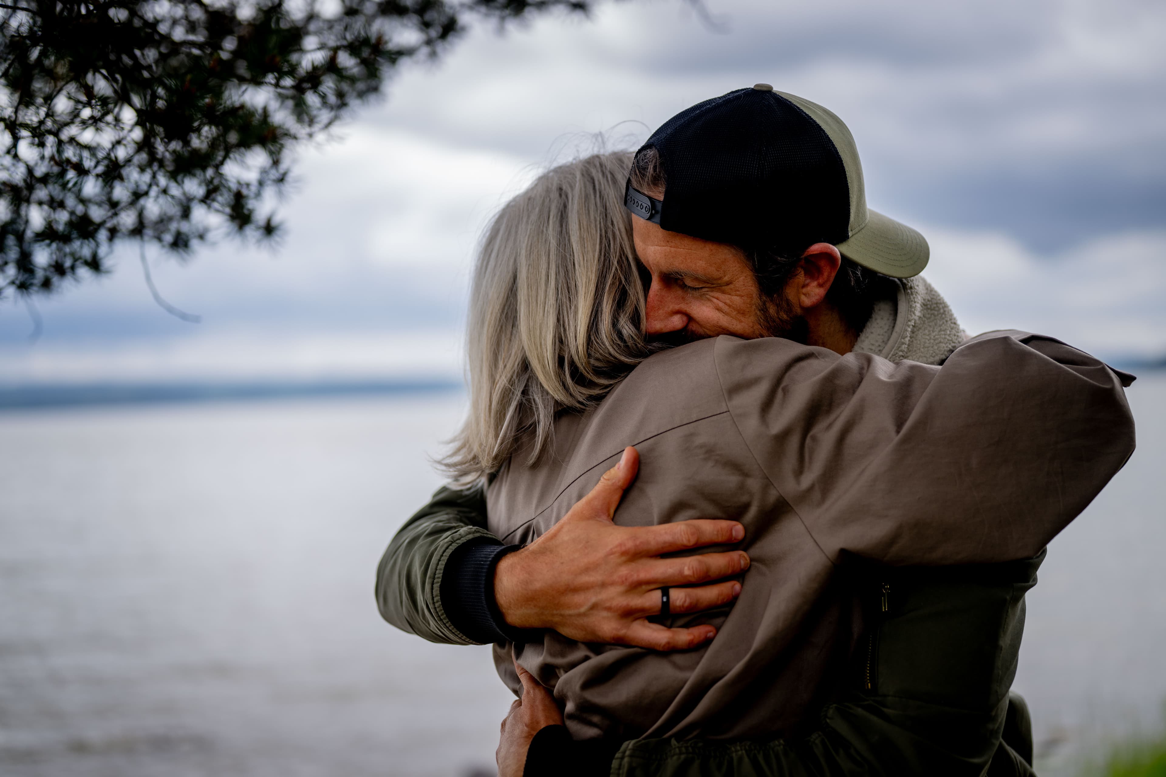 En mann med caps klemmer varmt om en eldre kvinne med grått hår ved et vann under en overskyet himmel, og begge virker glade og fornøyde.